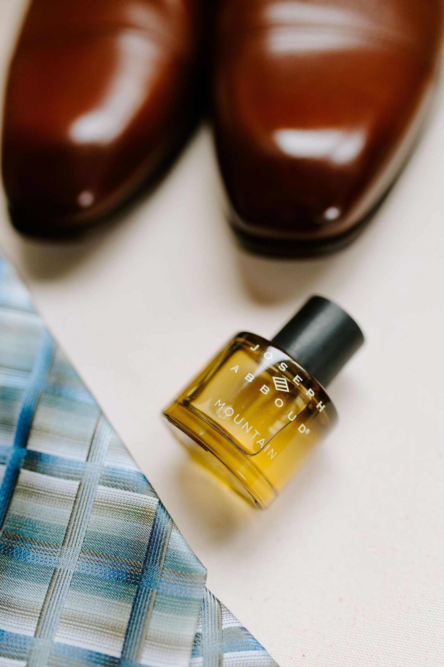 Elegant brown leather dress shoes and amber perfume bottle arranged with a blue patterned fabric on white surface.