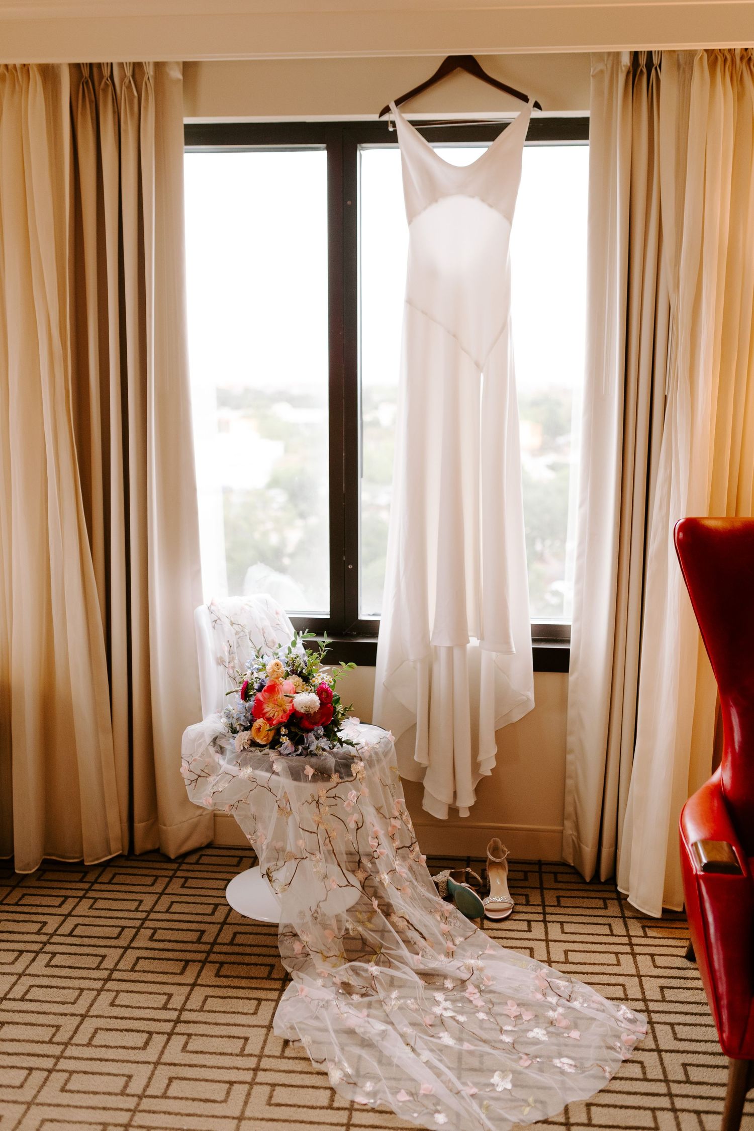 Elegant white wedding dress hangs by window with lace veil and floral bouquet arranged on patterned carpet below.