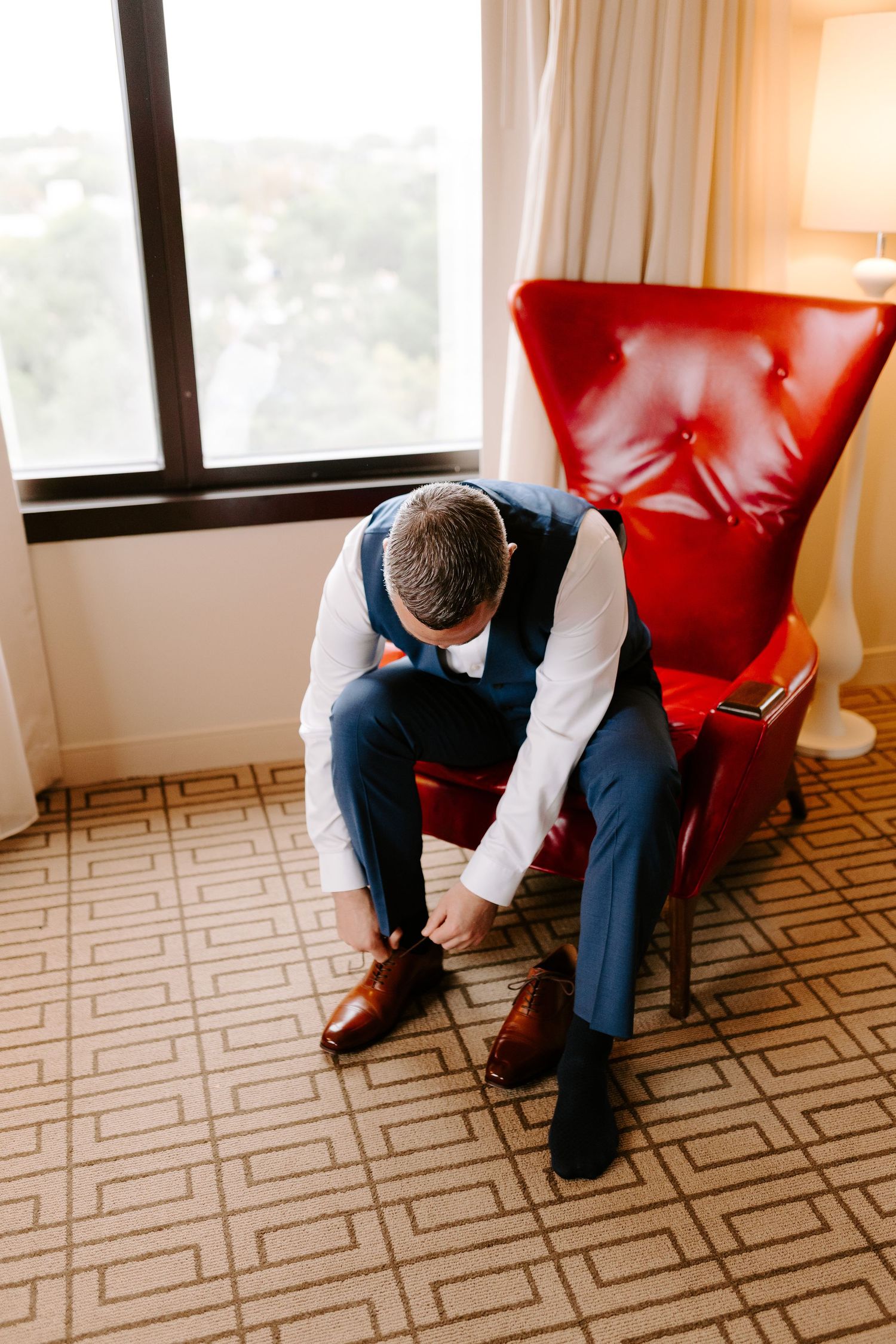 Someone in formal attire sits hunched over on a red leather chair while adjusting their brown dress shoes on patterned carpet.