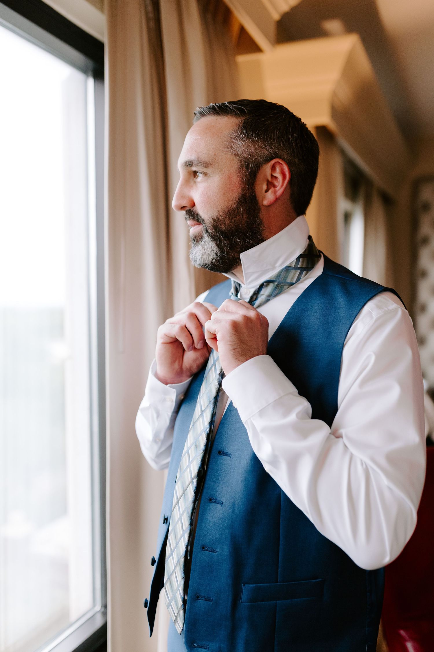 Man in navy blue vest and white dress shirt adjusts his tie while looking out a window.