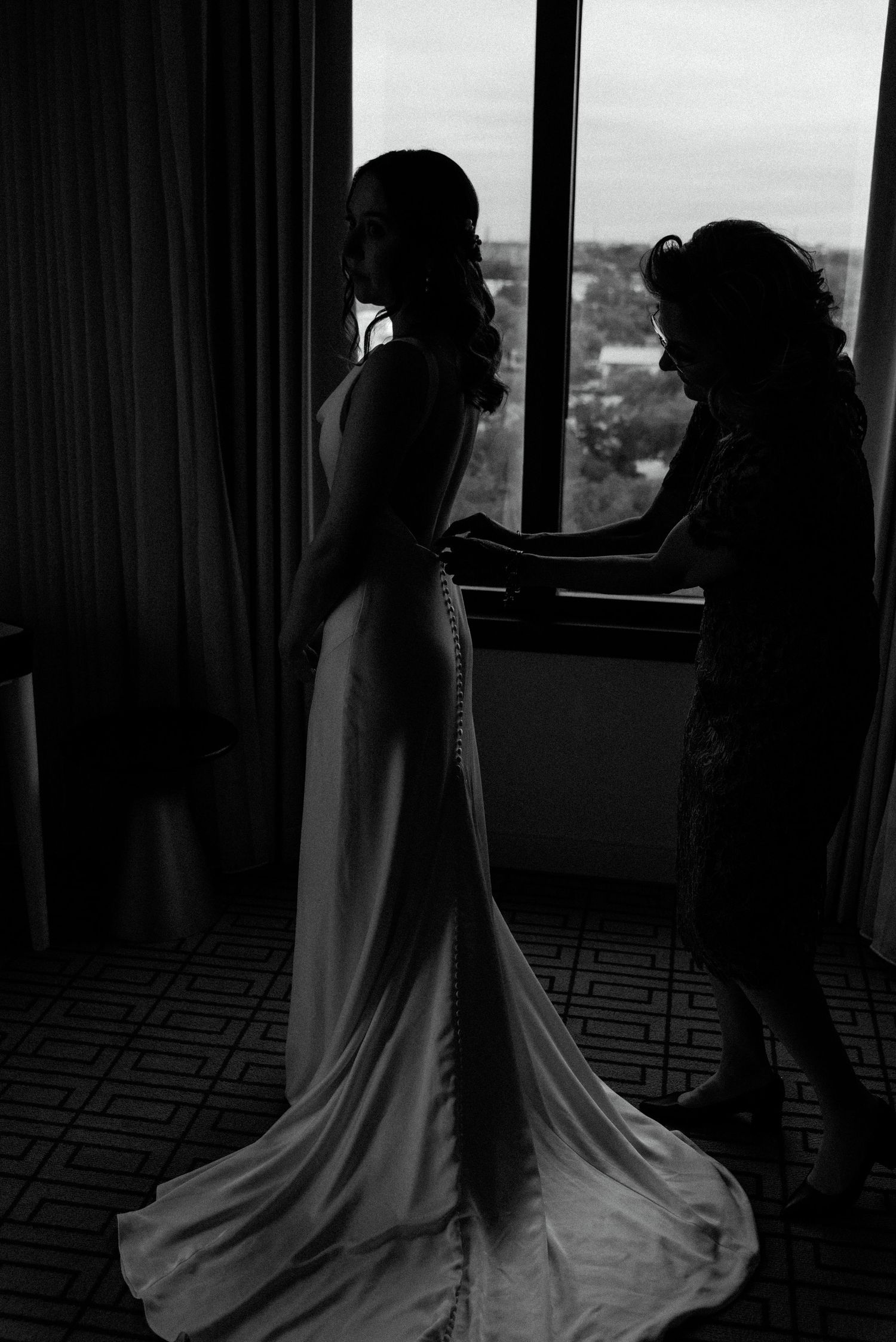 A black and white silhouette of final wedding dress adjustments being made near a window with natural light streaming in.
