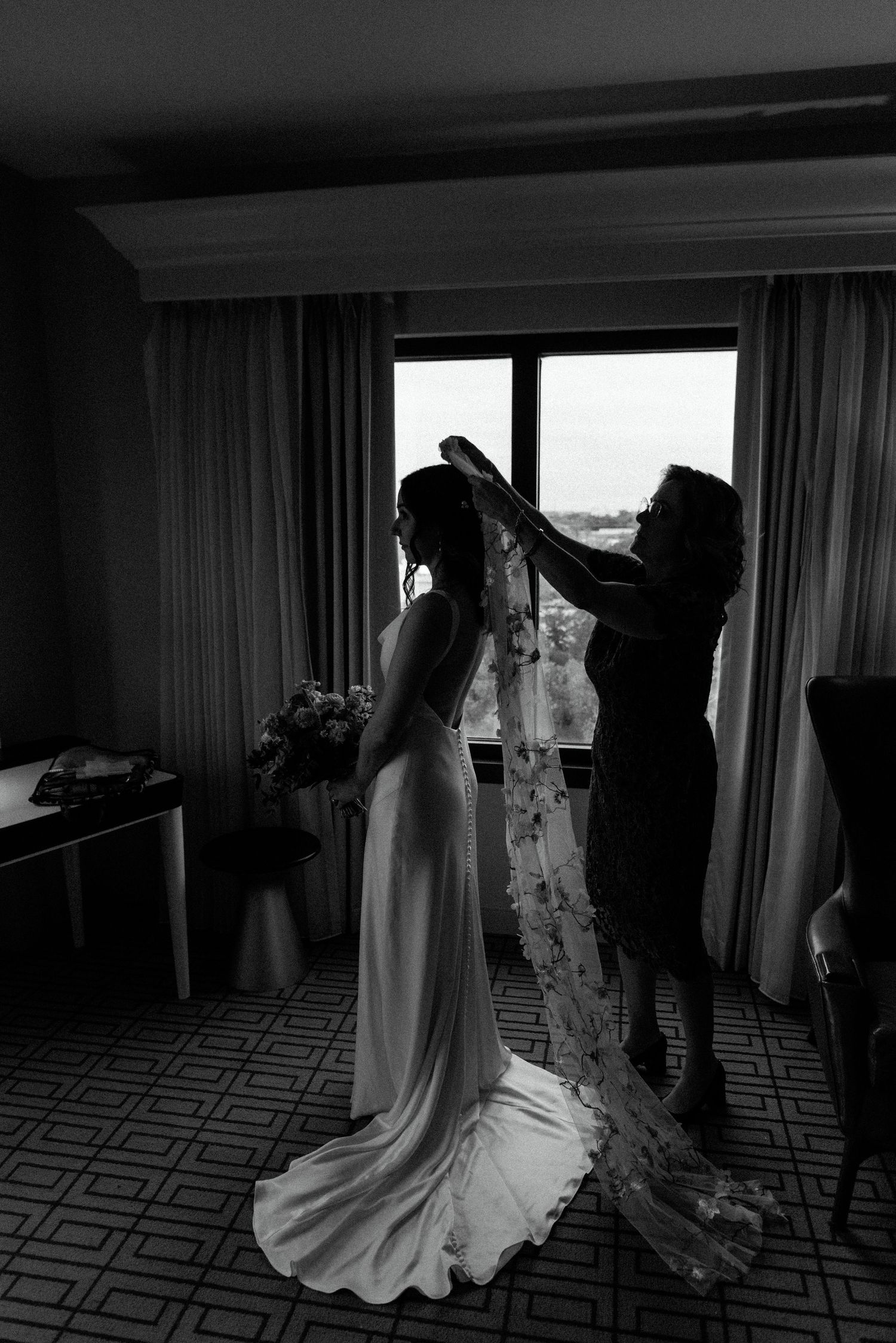 Black and white image of wedding dress preparations with veil being adjusted near a window.