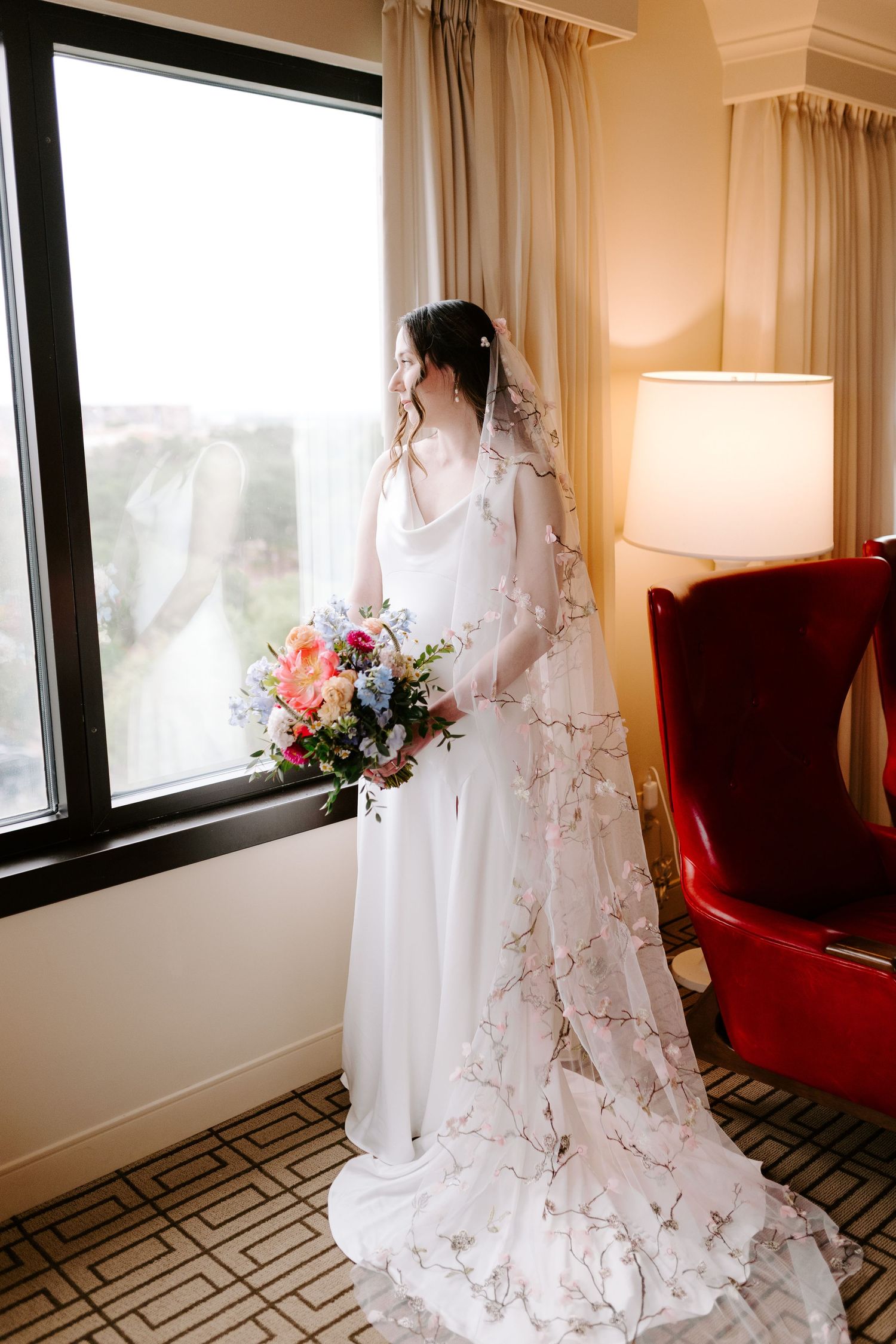 A bride in a flowing white lace veil and gown holds a vibrant bouquet while standing by a window near a red armchair.