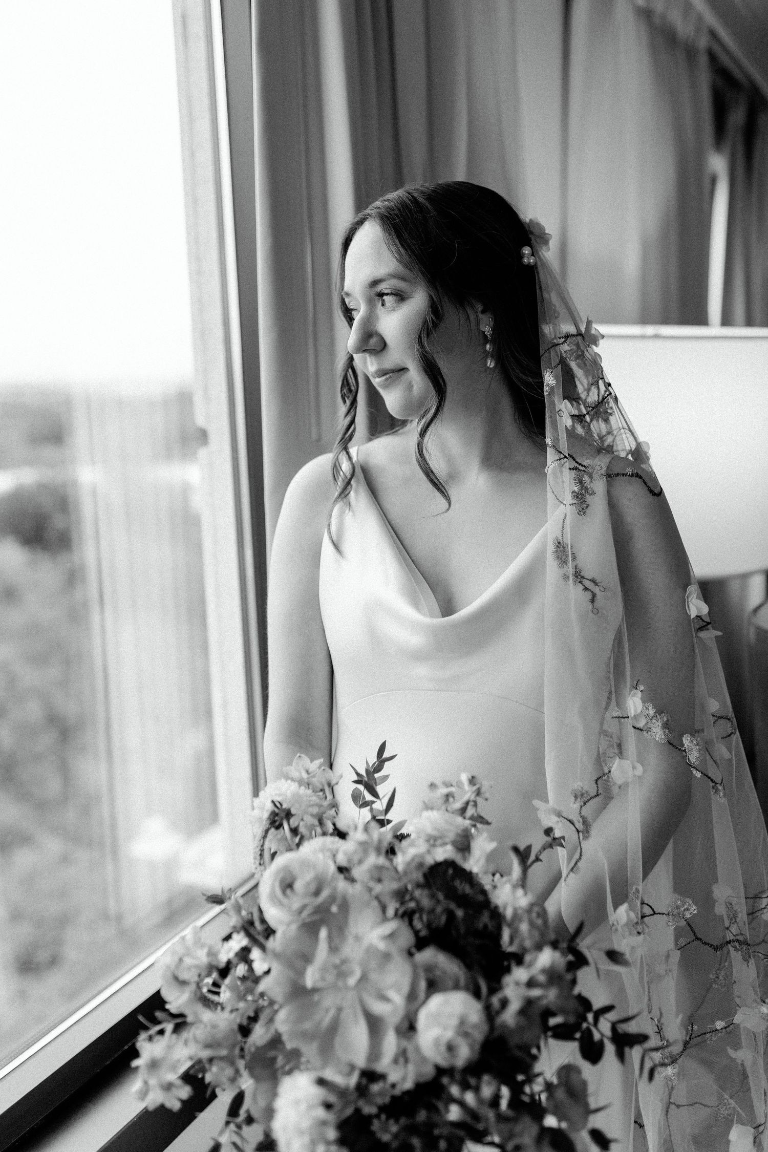 Elegant wedding gown and bouquet details captured in black and white by a window.