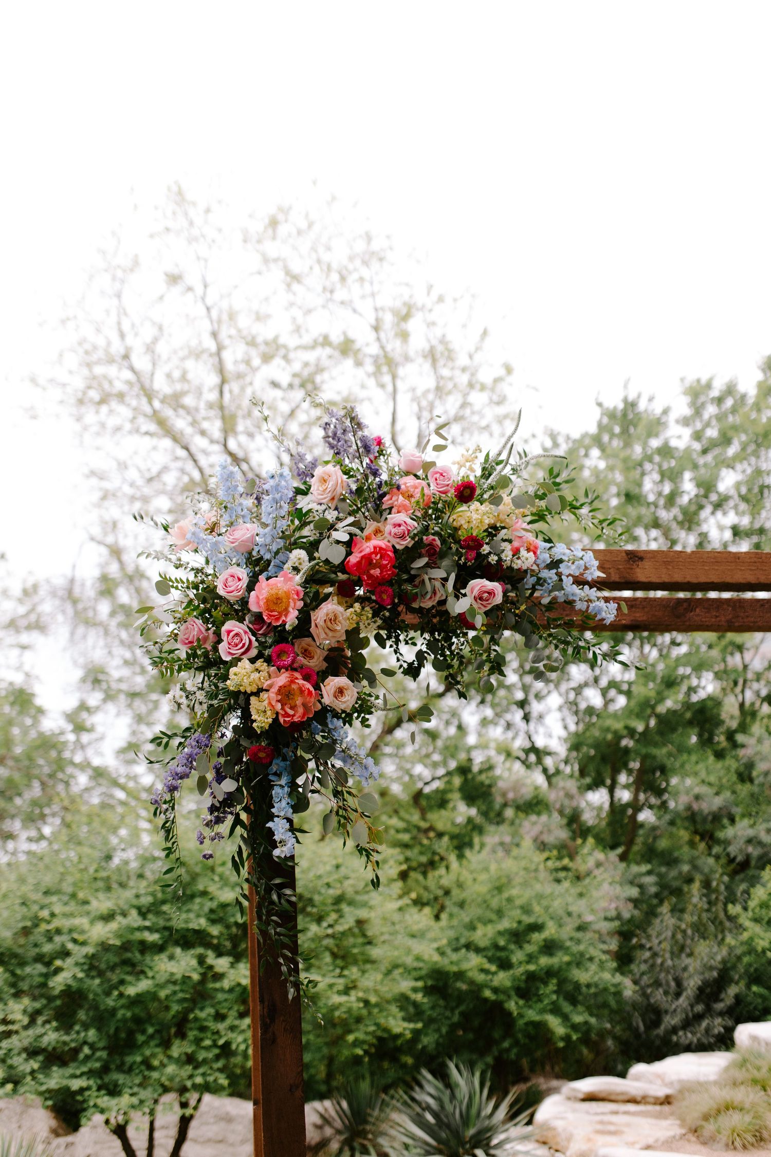 Beautiful floral arrangement with pink and coral roses adorns a wooden arch at an outdoor wedding ceremony setting.
