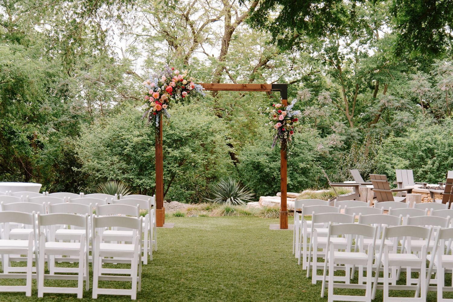 Outdoor wedding ceremony setup with wooden arch decorated with flowers and white chairs on a green lawn.