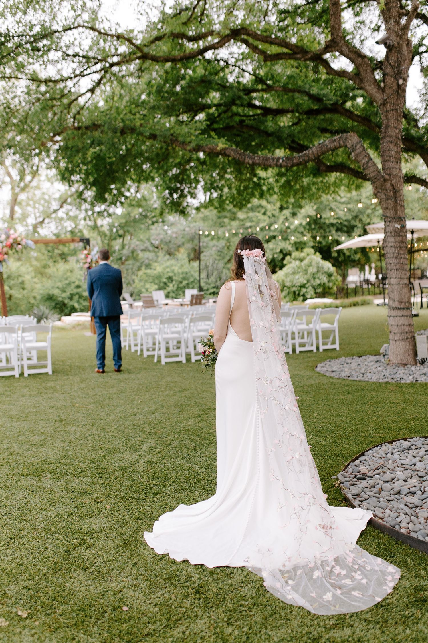 Wedding ceremony setup in a garden with white chairs and floral arch under large trees.