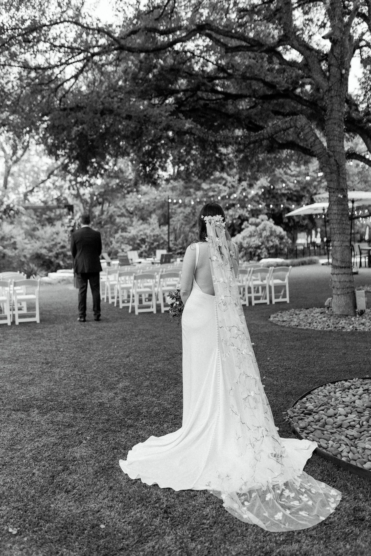 A black and white photo of a bride in a lace-detailed wedding dress with cathedral veil during first look.
