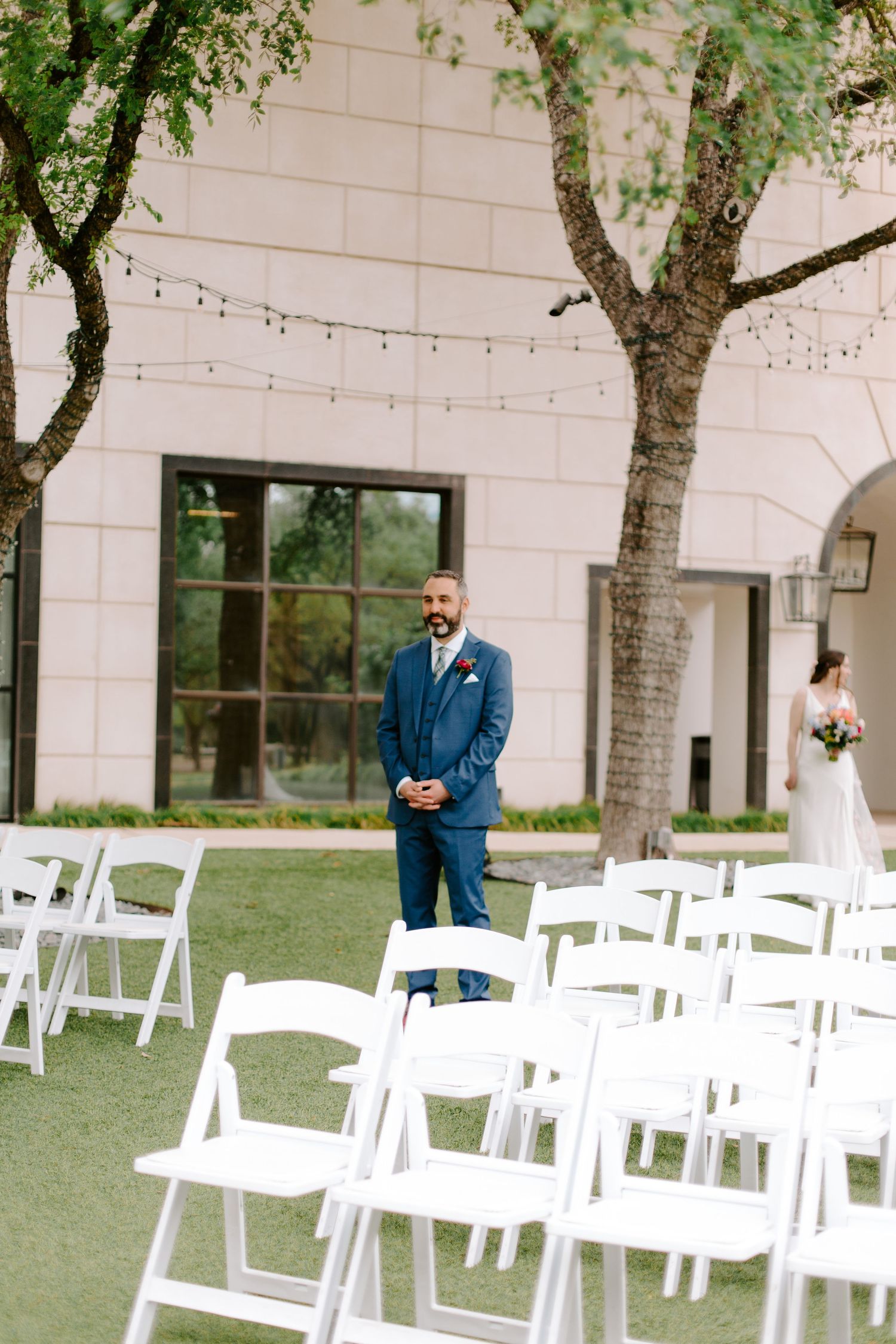 An outdoor wedding ceremony setup with white chairs and a groom in navy suit waiting at the altar.