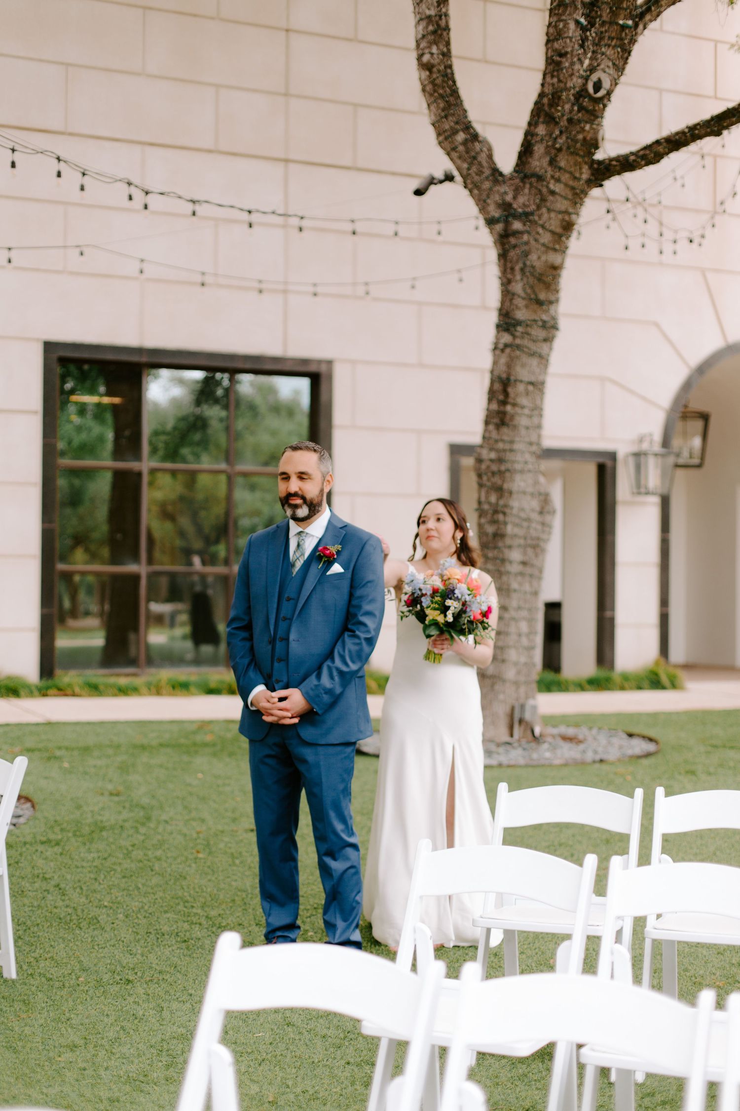 A bride holding a bouquet approaches her groom during their first look at an outdoor wedding venue.