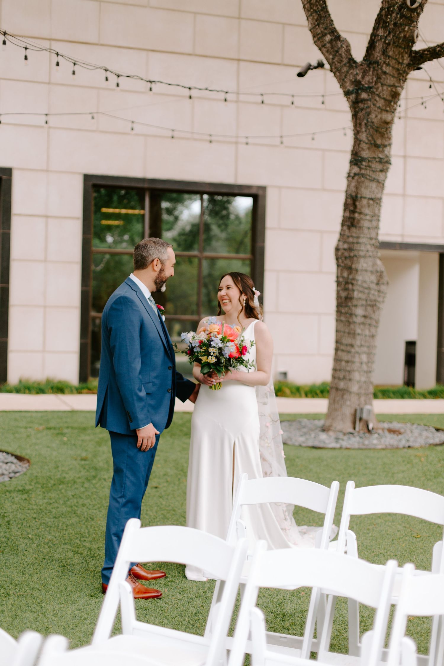 An outdoor wedding ceremony setup with white chairs and string lights hanging between trees.