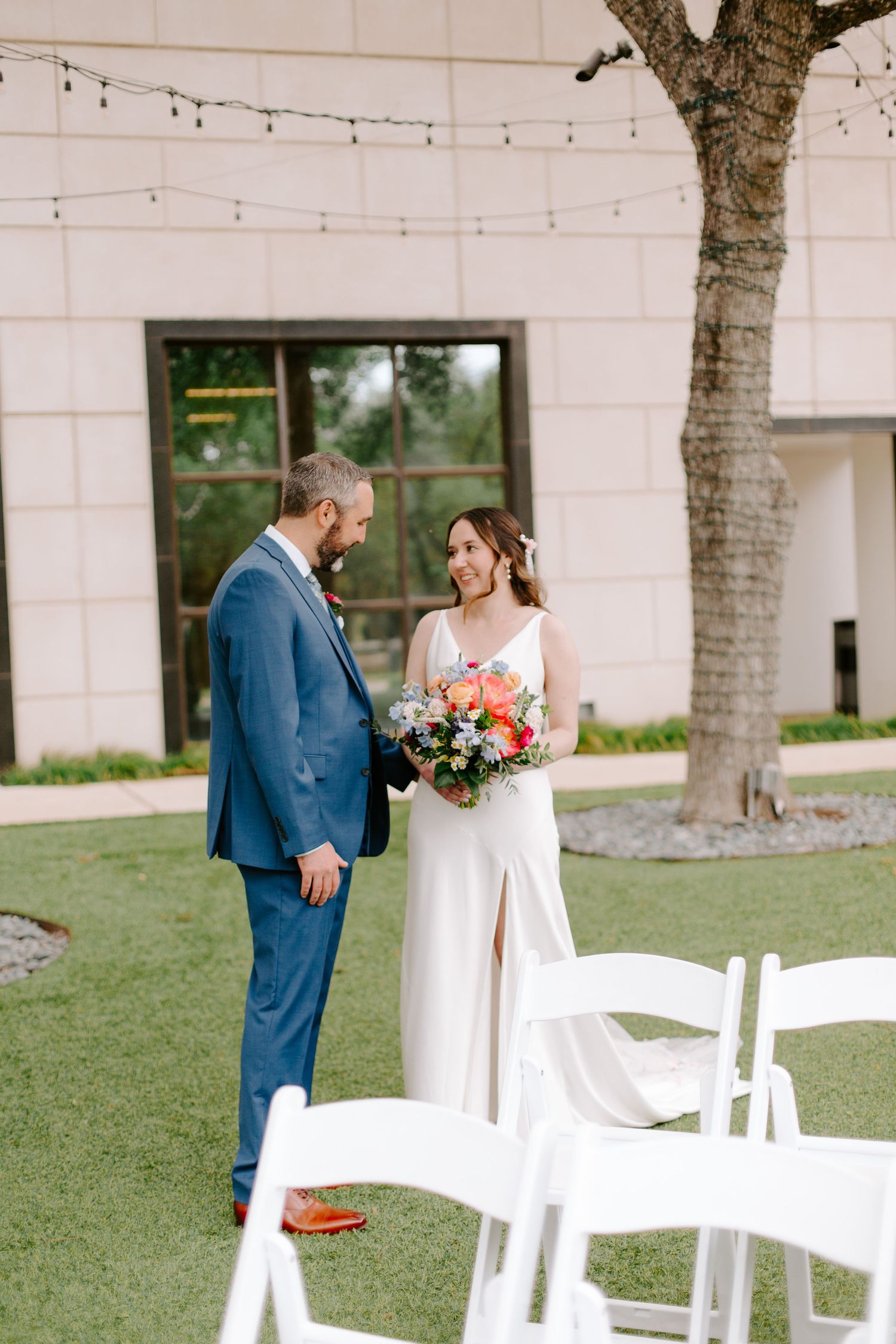 A couple shares a tender moment at their outdoor wedding, standing on grass near white chairs with string lights overhead.