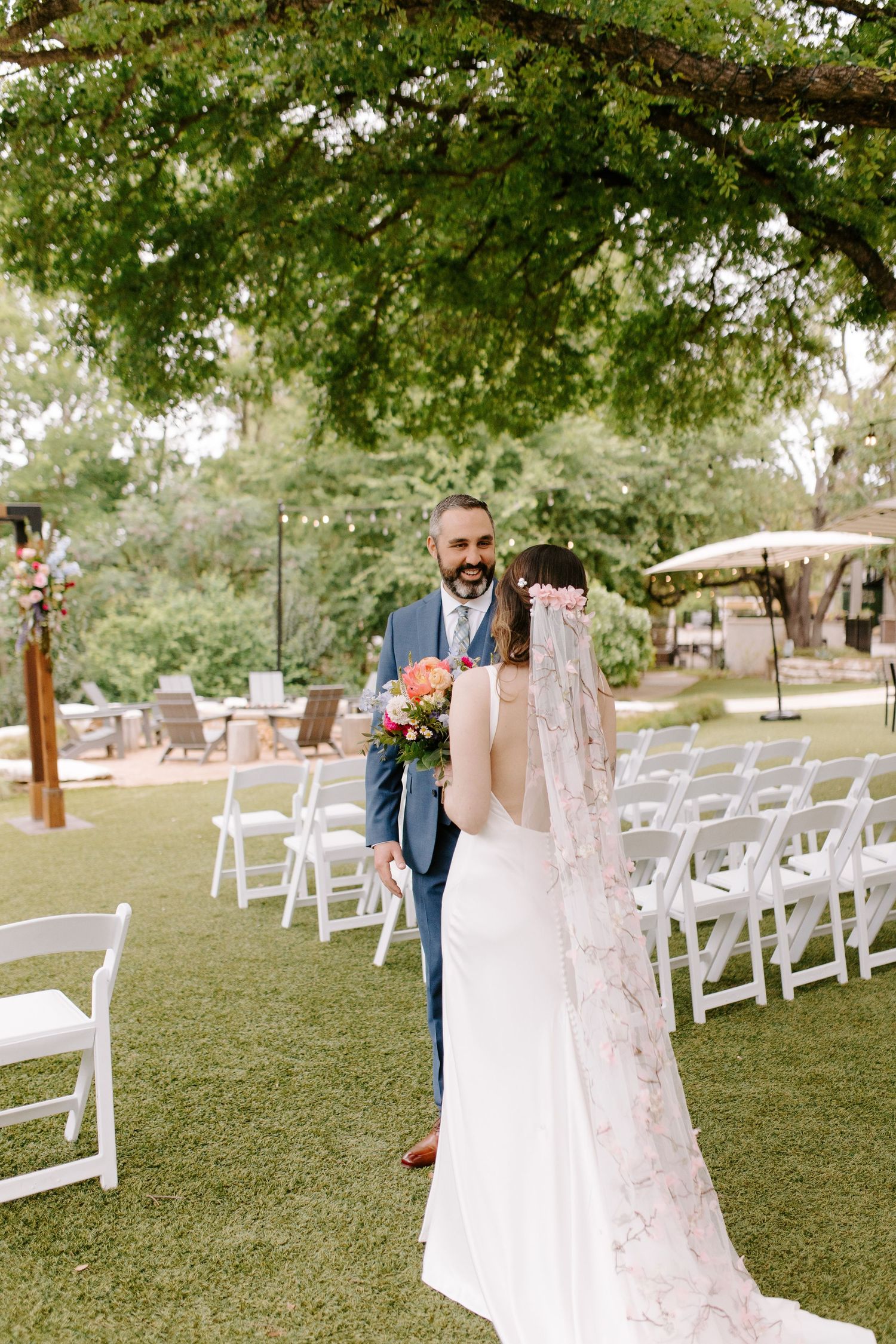 A wedding ceremony setup outdoors with white chairs on a grass lawn under trees with two people embracing.