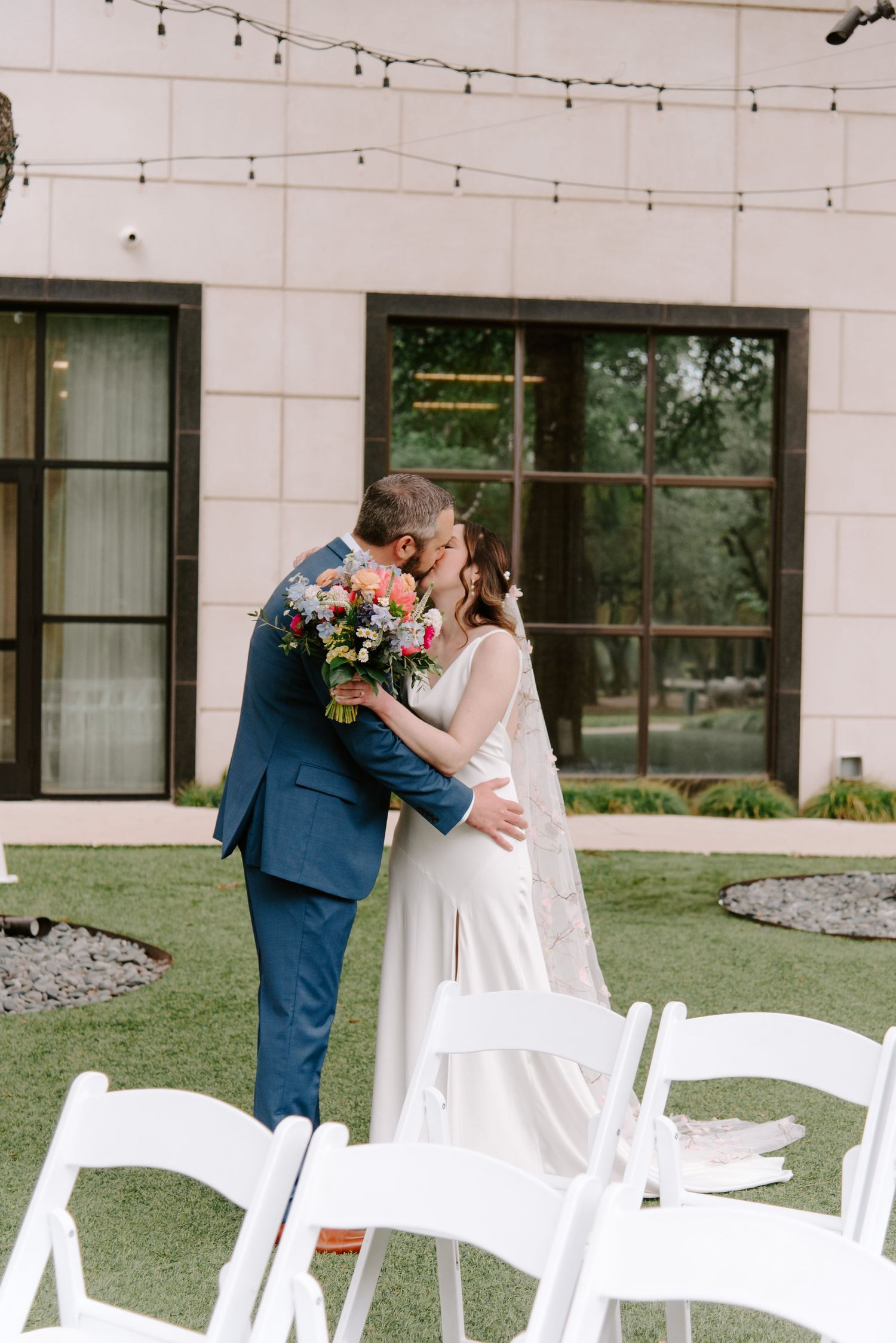 A romantic wedding moment outdoors with white chairs, string lights, and a colorful bouquet against a modern venue wall.