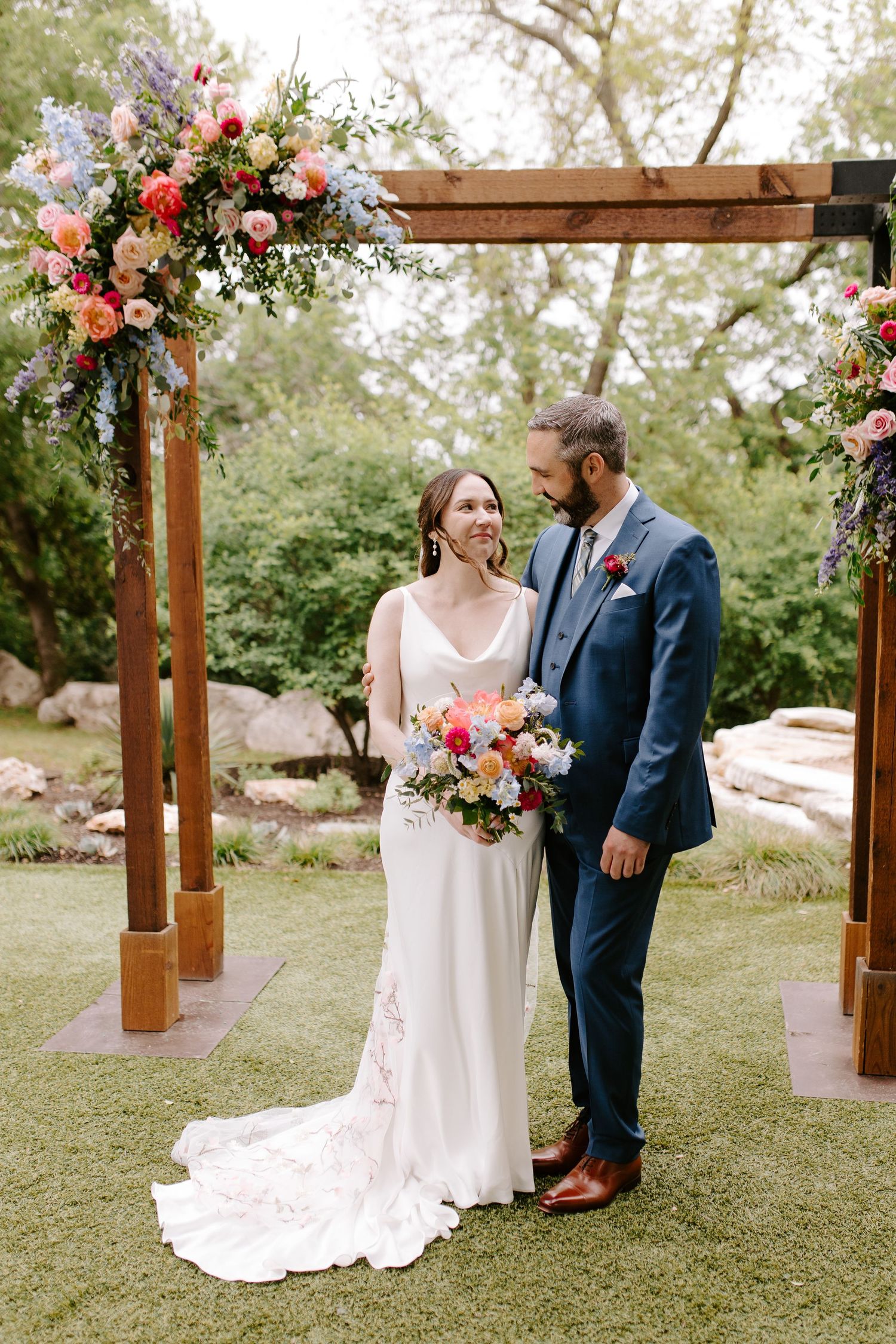 A wedding couple stands beneath a wooden arbor adorned with vibrant coral and pink floral arrangements in a garden setting.