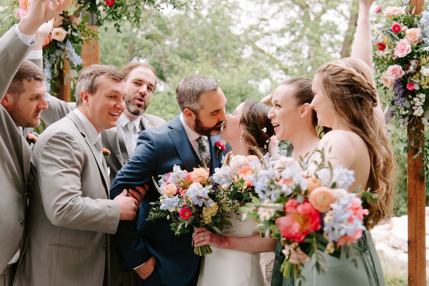 Wedding party laughing joyfully together under floral arch with pink and peach bouquets.