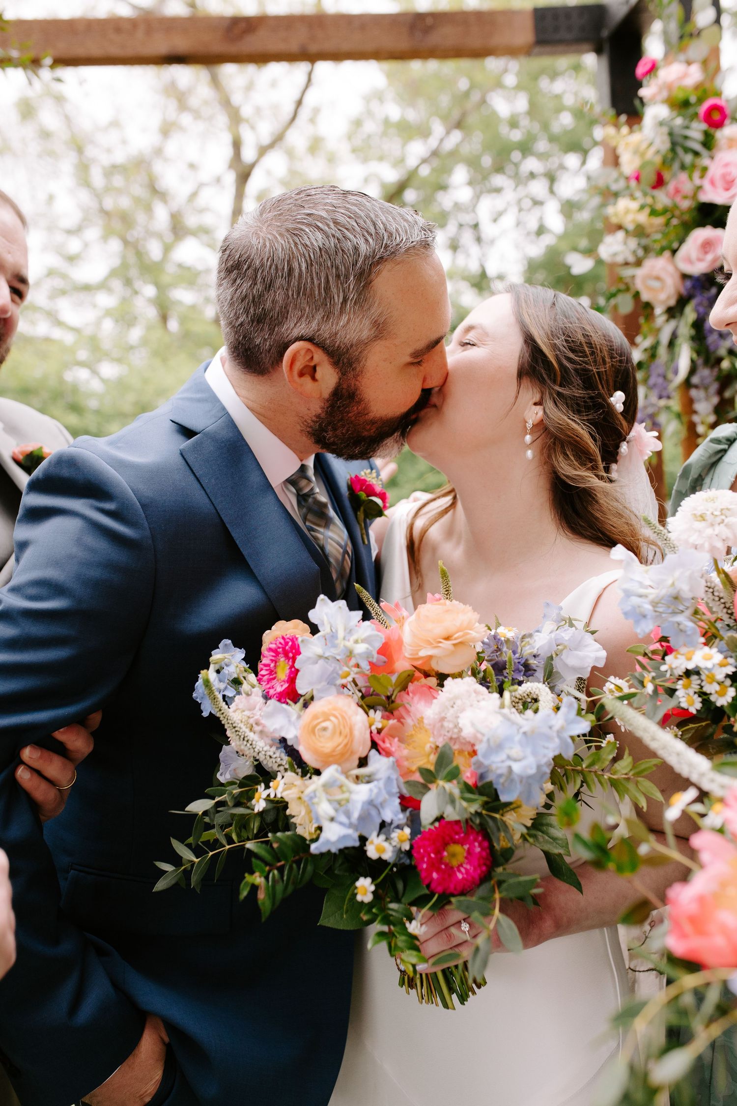 A couple shares a romantic kiss at their wedding surrounded by vibrant floral arrangements.