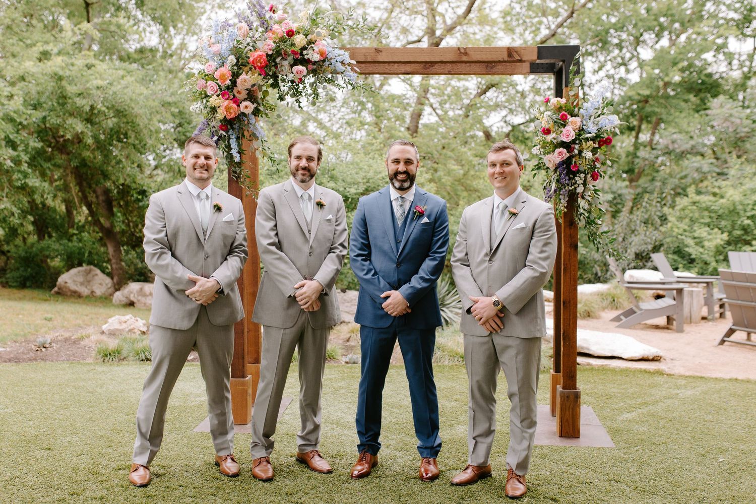 Wedding party stands beneath wooden arch decorated with pink and orange flowers in an outdoor garden setting.