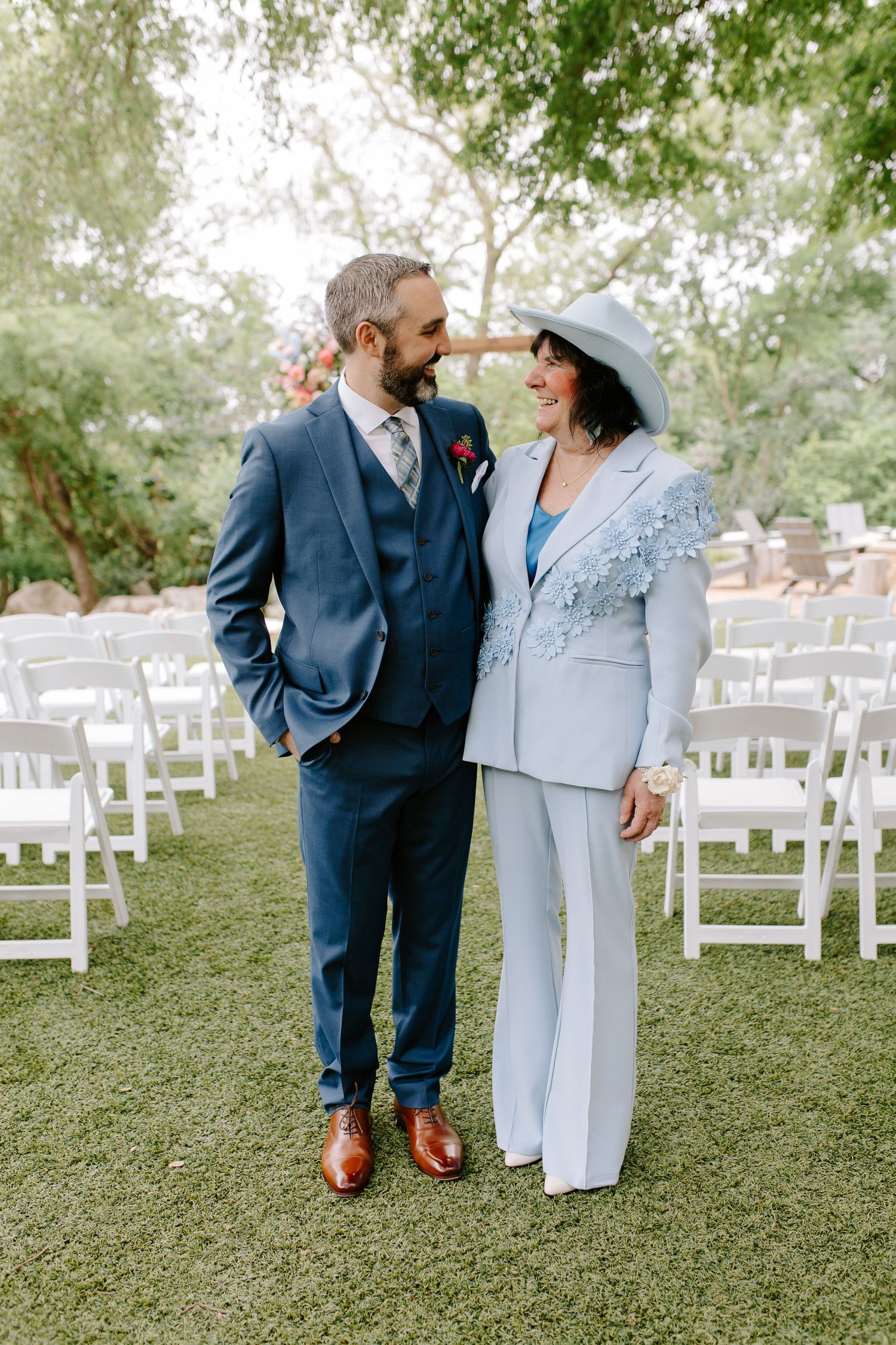 Two people in formal attire stand together on a grass lawn with white chairs arranged for a wedding ceremony.