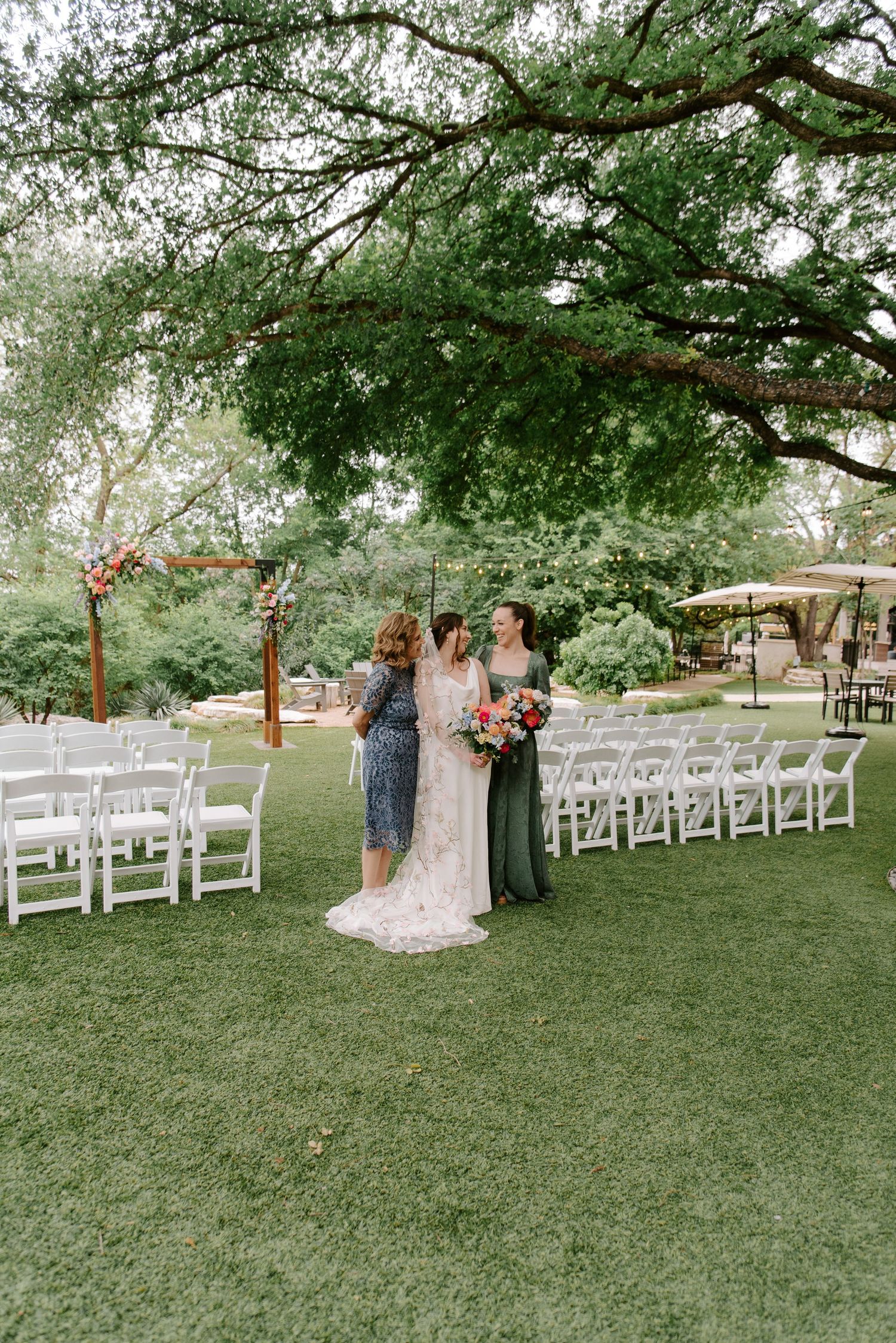 Beautiful outdoor wedding ceremony setup under a large tree with white chairs and floral arch decorations.
