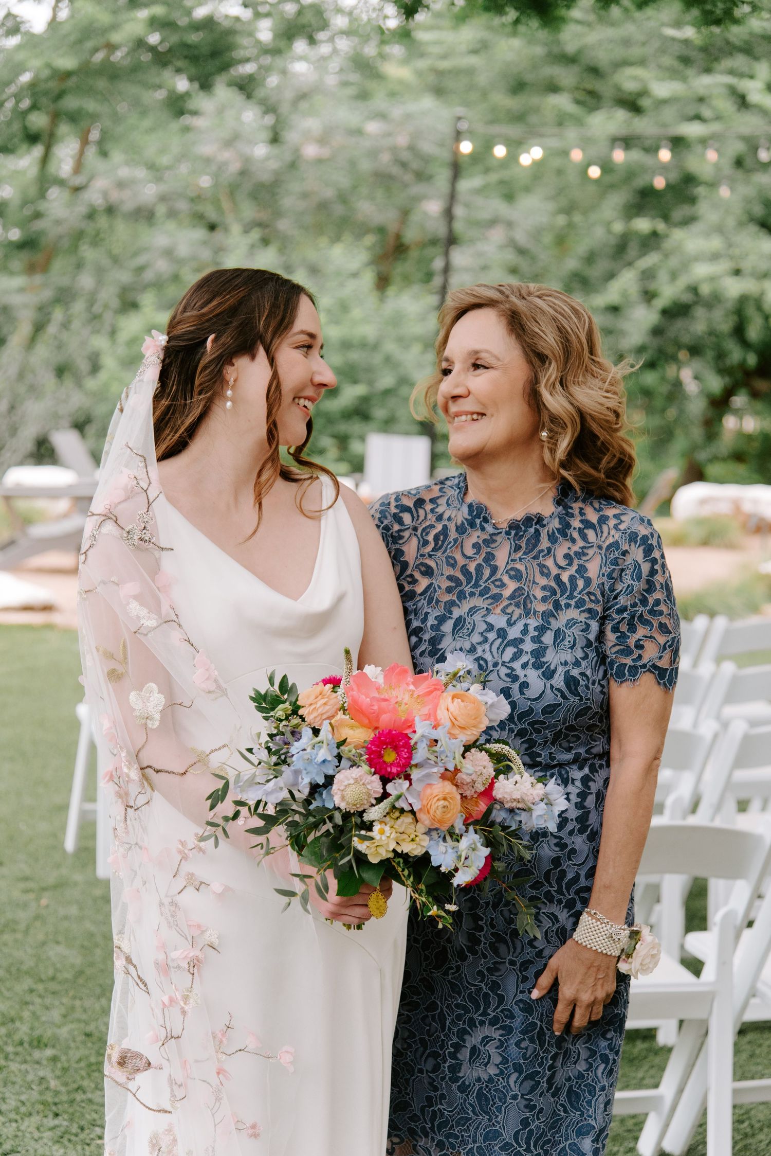 Two women share a joyful moment at a wedding, one in a white gown and another in a blue floral dress.