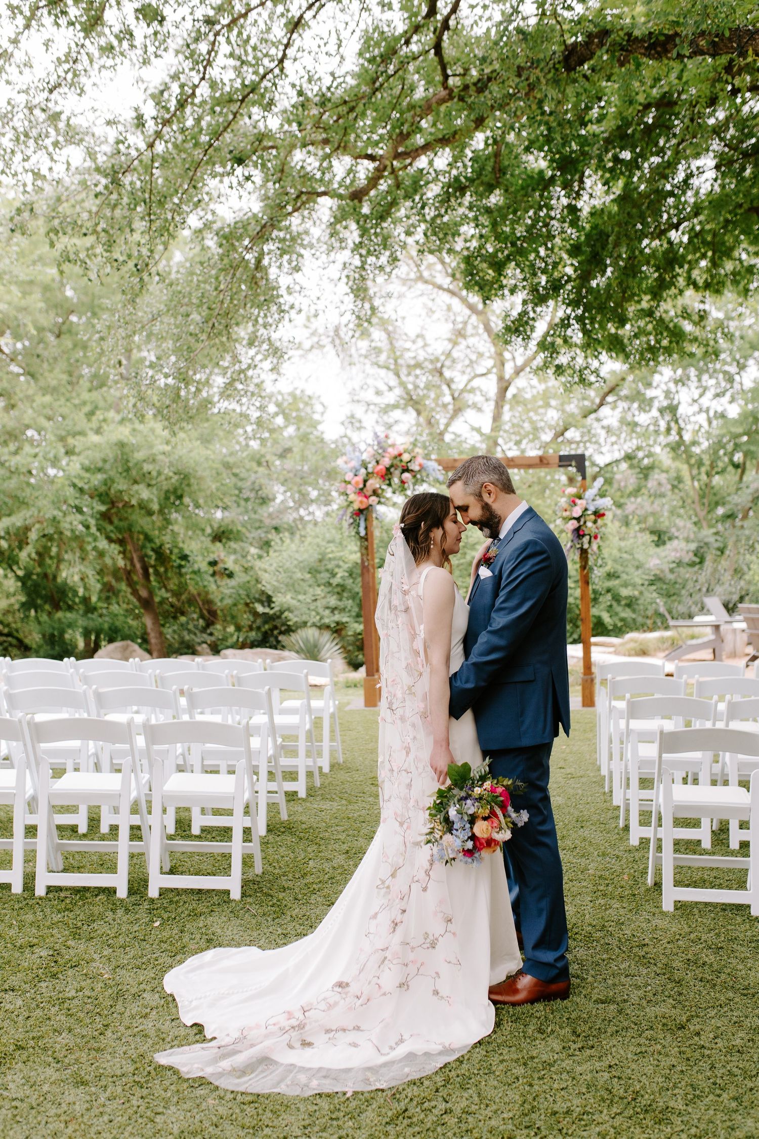 Romantic outdoor wedding photos with white chairs and wooden arch under trees.