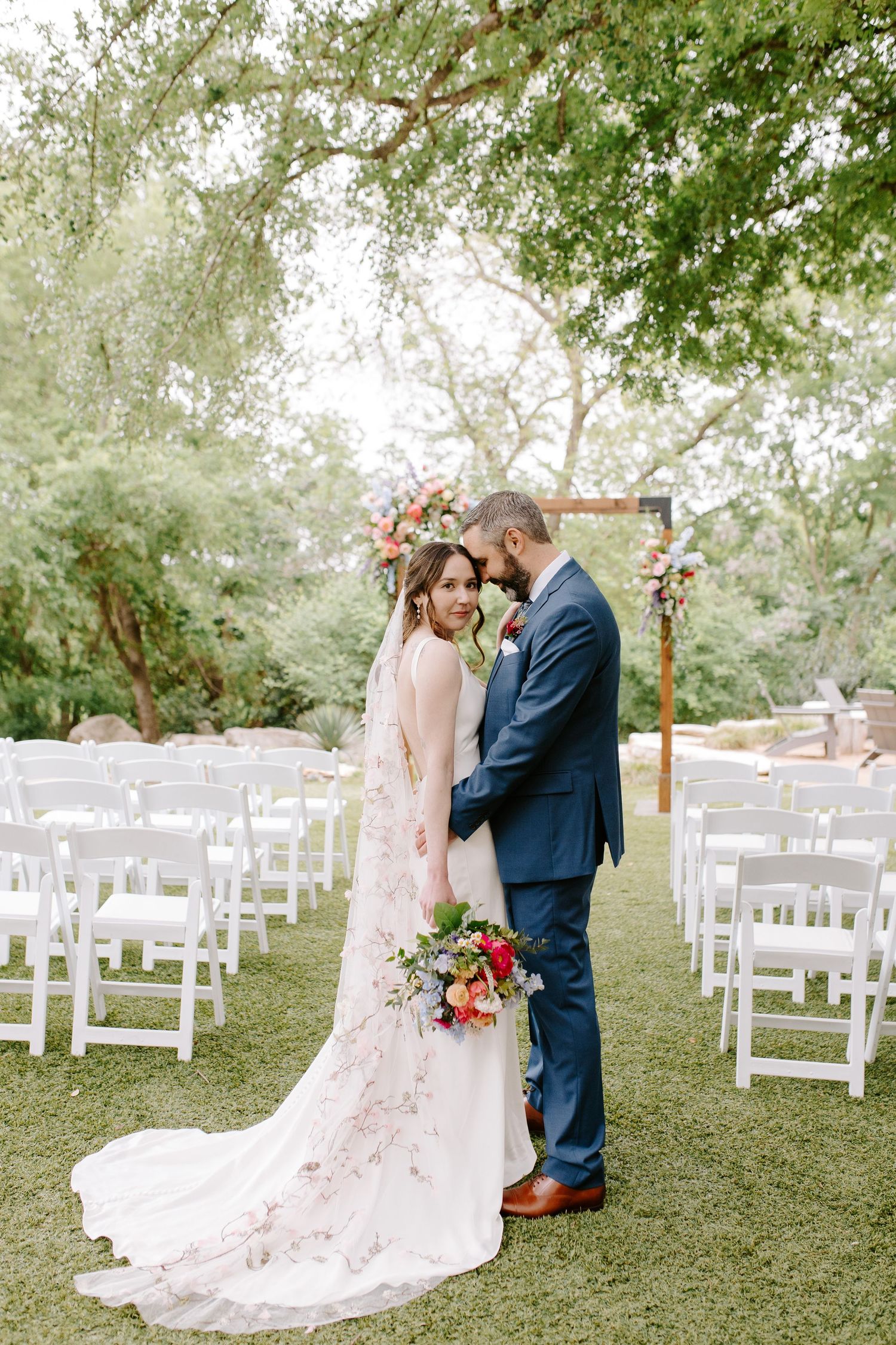 Wedding couple posing in a garden setting with white chairs and floral arrangements.