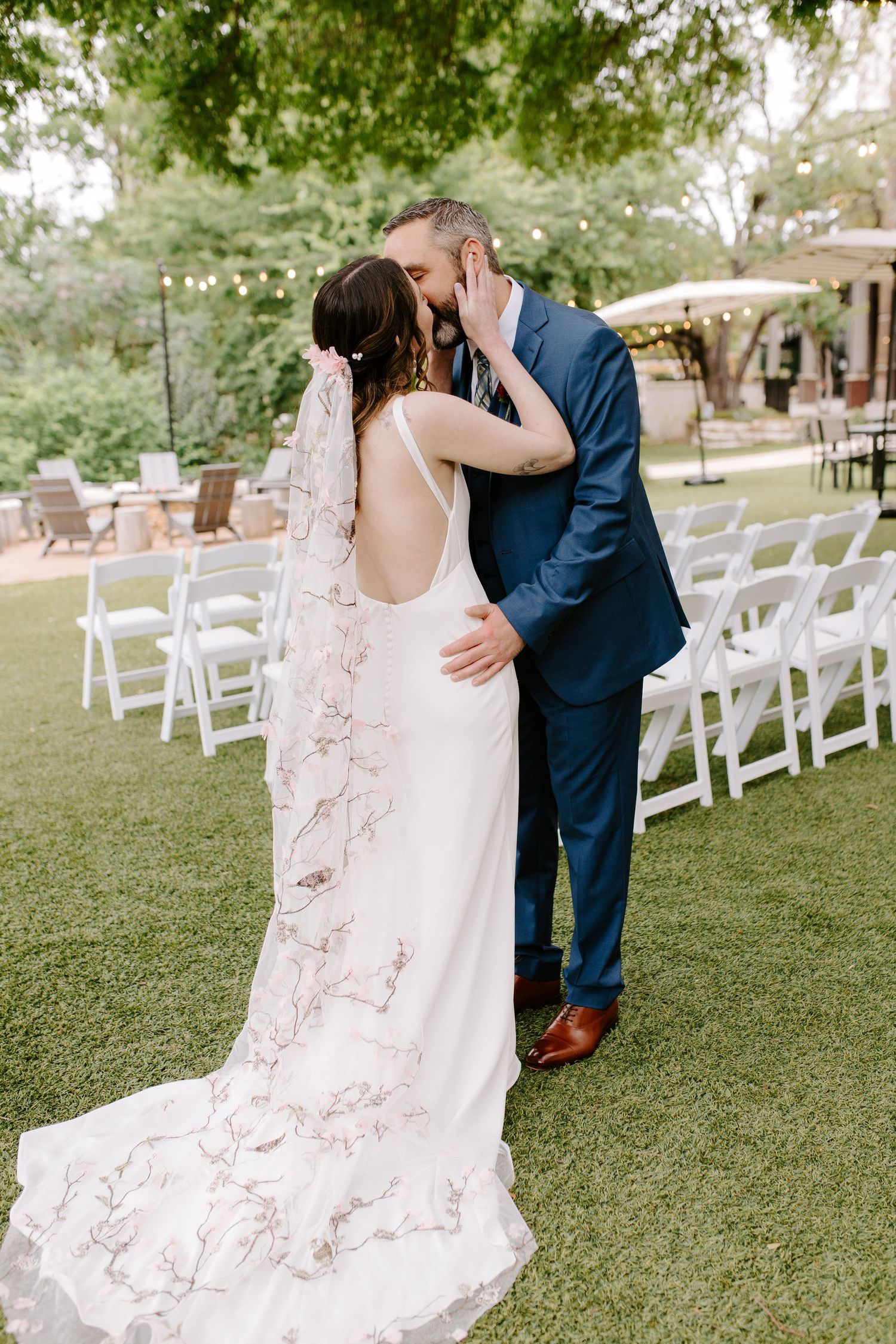 A romantic wedding moment outdoors with a couple embracing on a manicured lawn with white chairs and string lights.