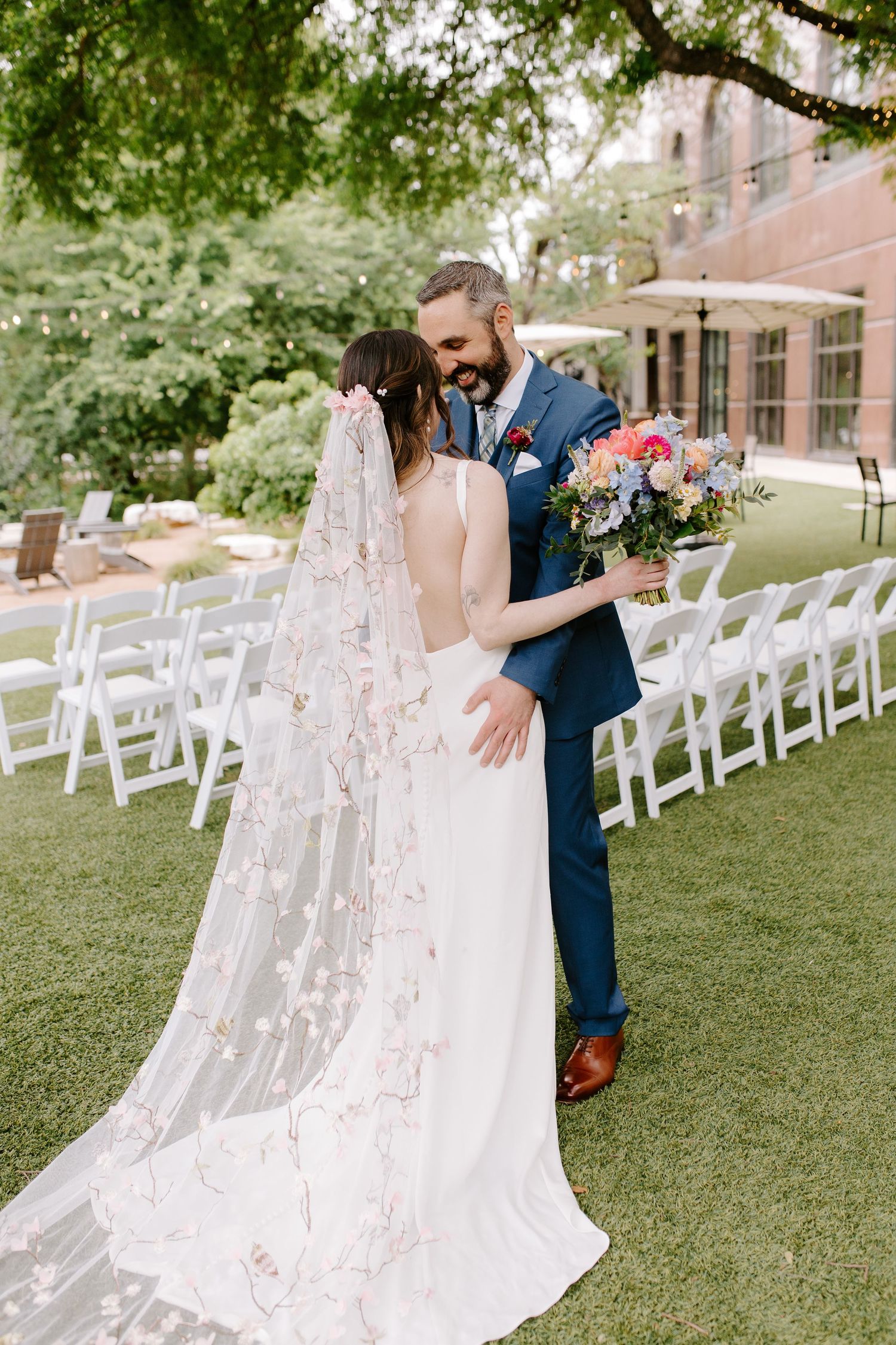 An intimate moment at an outdoor wedding with a stunning backless gown and delicate floral veil.