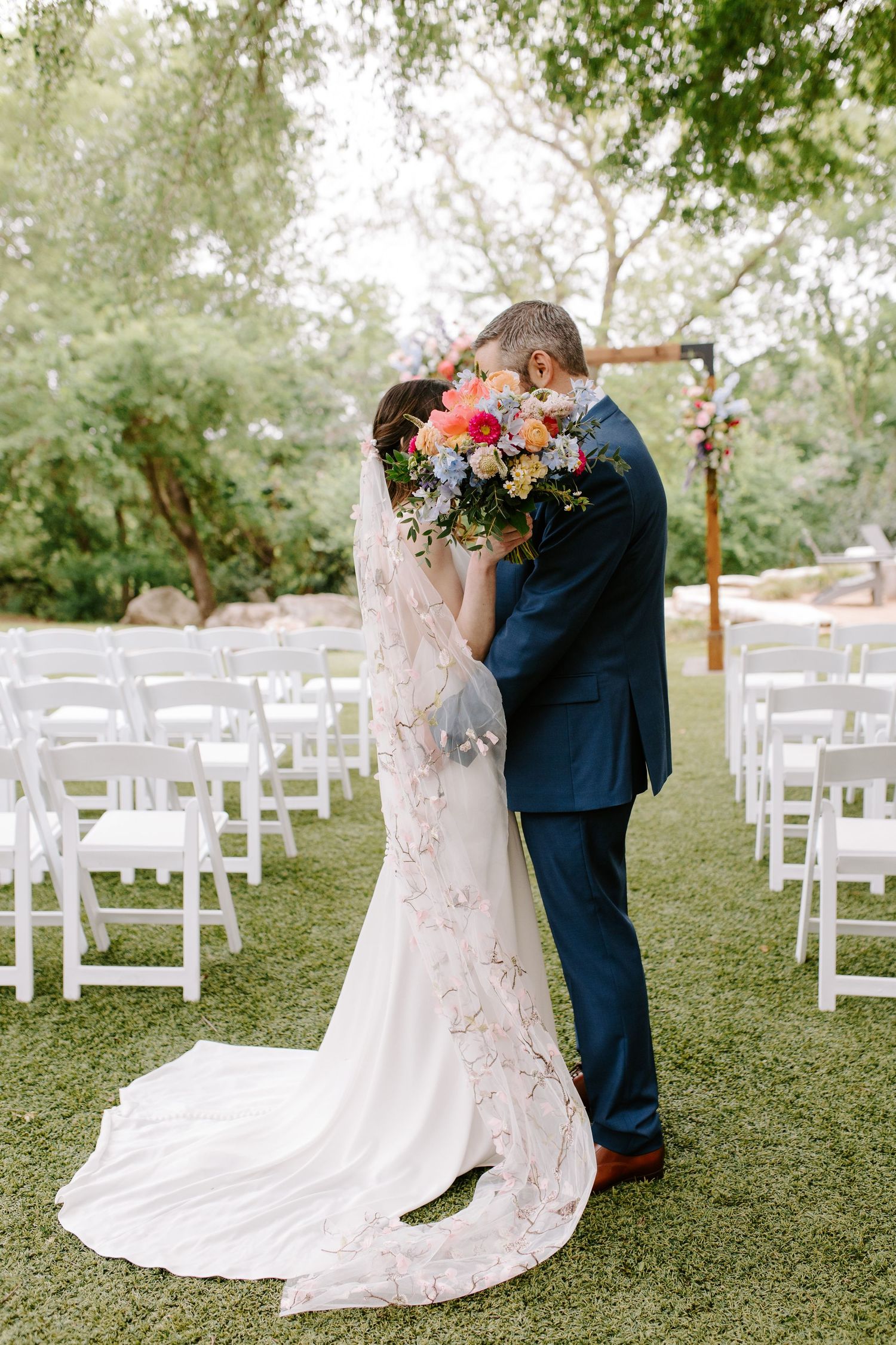 A romantic moment under a rustic wooden arch with white chairs and colorful flowers at an outdoor wedding ceremony.