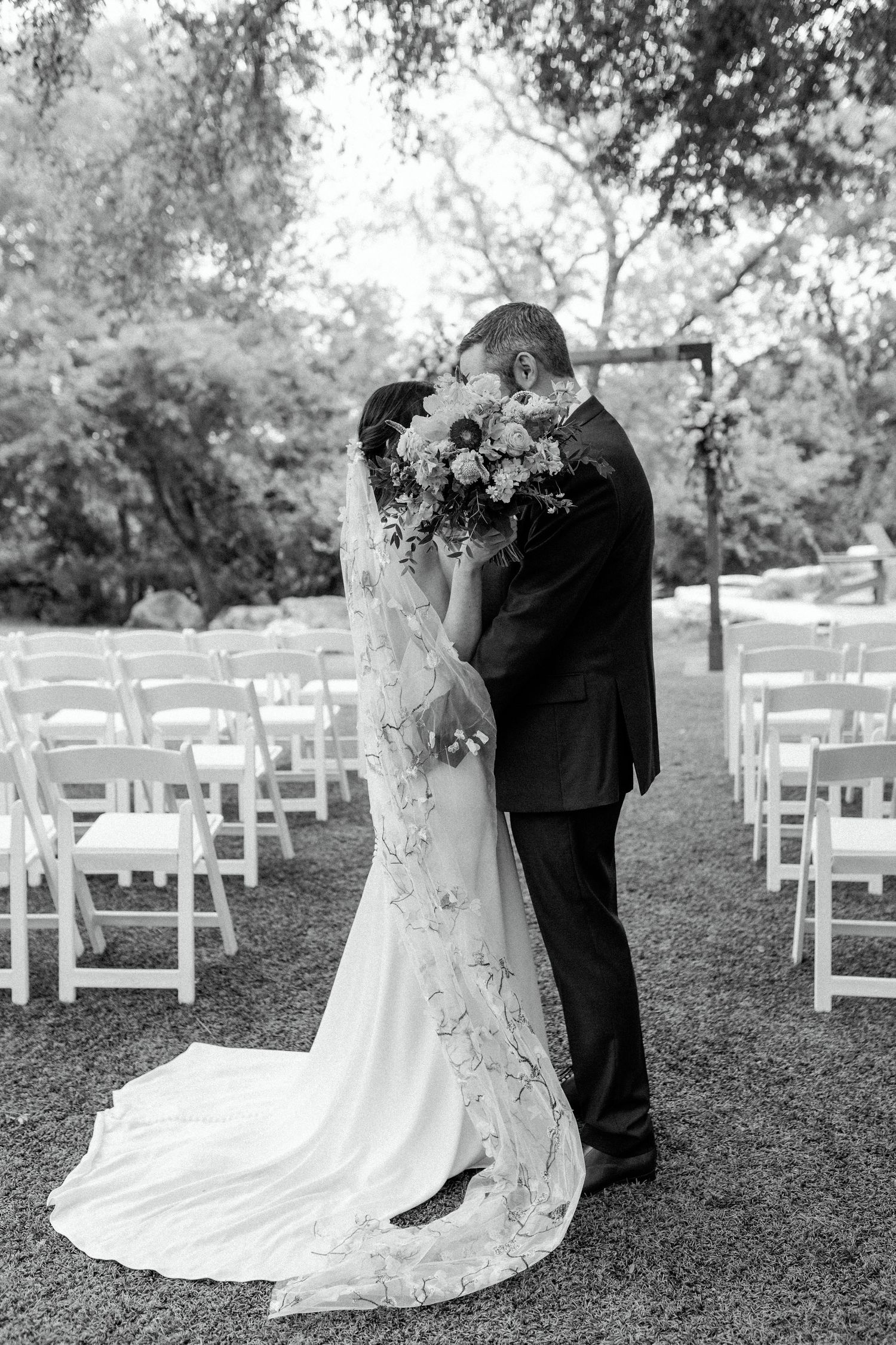 Black and white photo of a wedding couple embracing at an outdoor ceremony with white chairs.