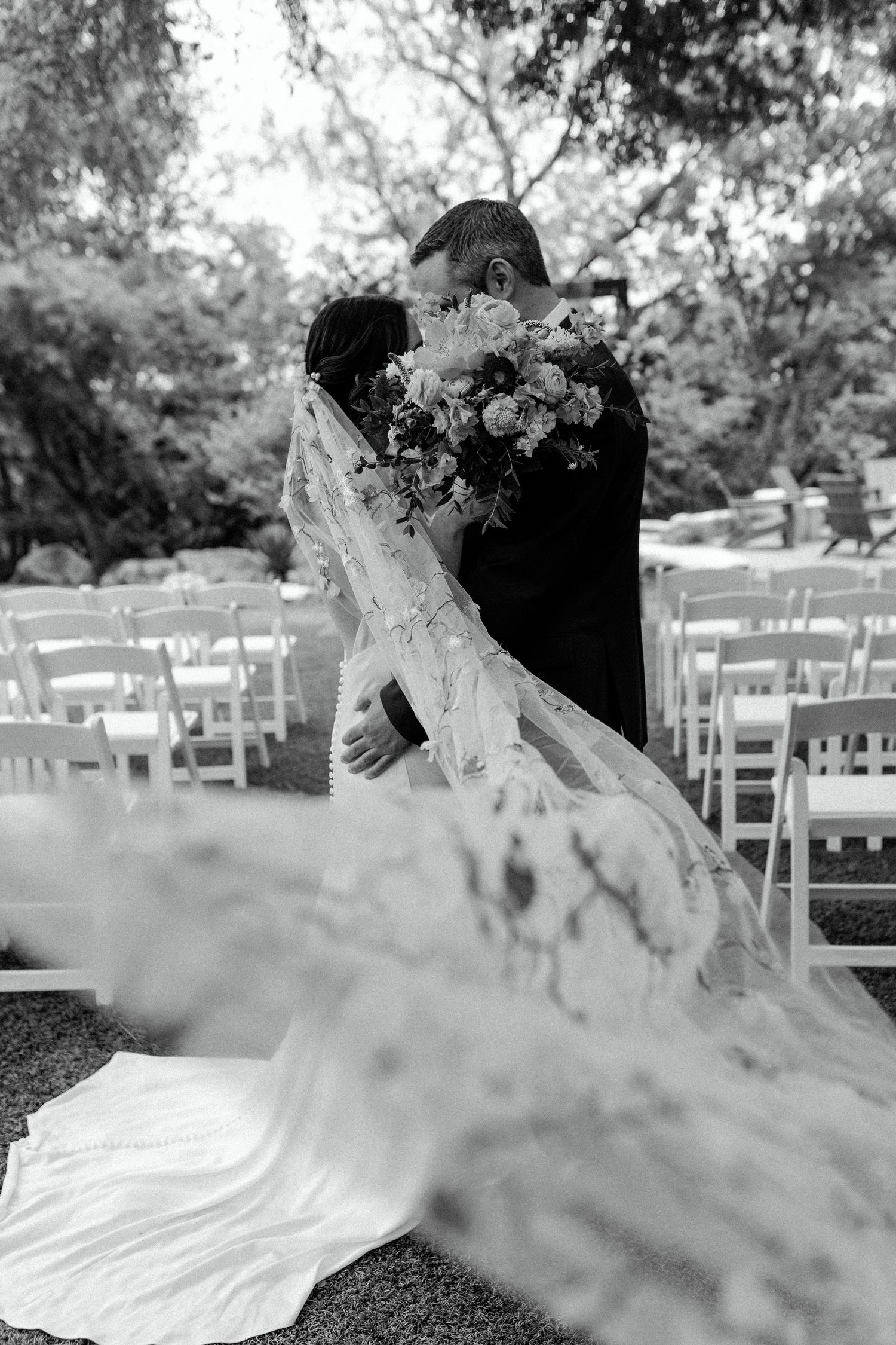 Artistic black and white wedding photo showing a couple embracing with the bride's lace dress flowing.
