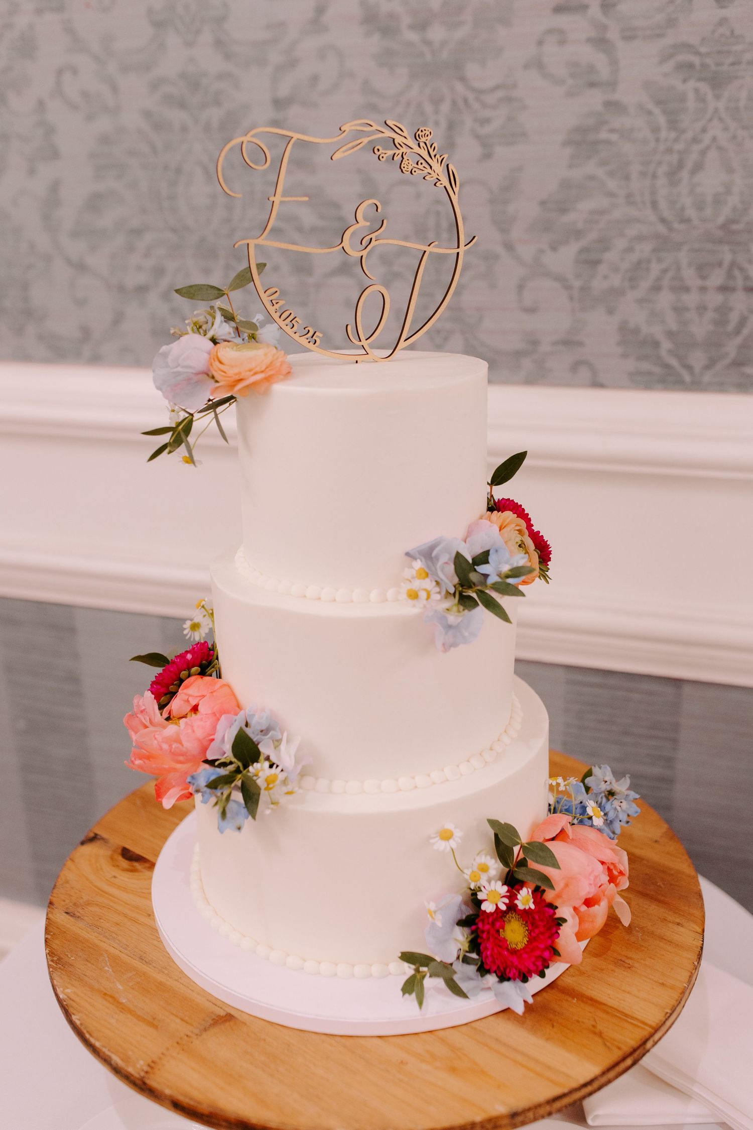Elegant white tiered wedding cake adorned with pink and red flowers, featuring a rose gold monogram topper on wooden base.