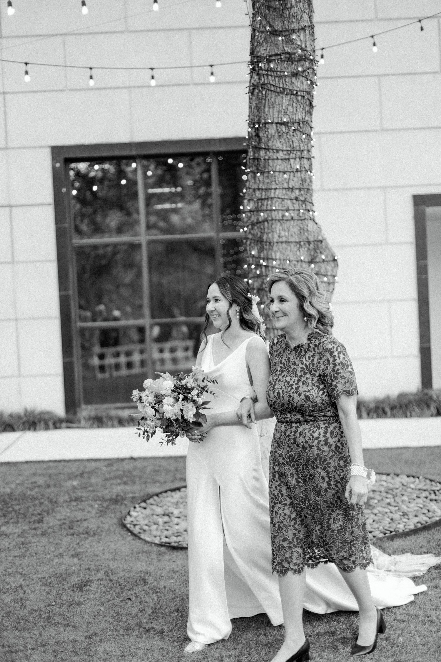 A black and white photo of two women walking together at an outdoor event with string lights visible.