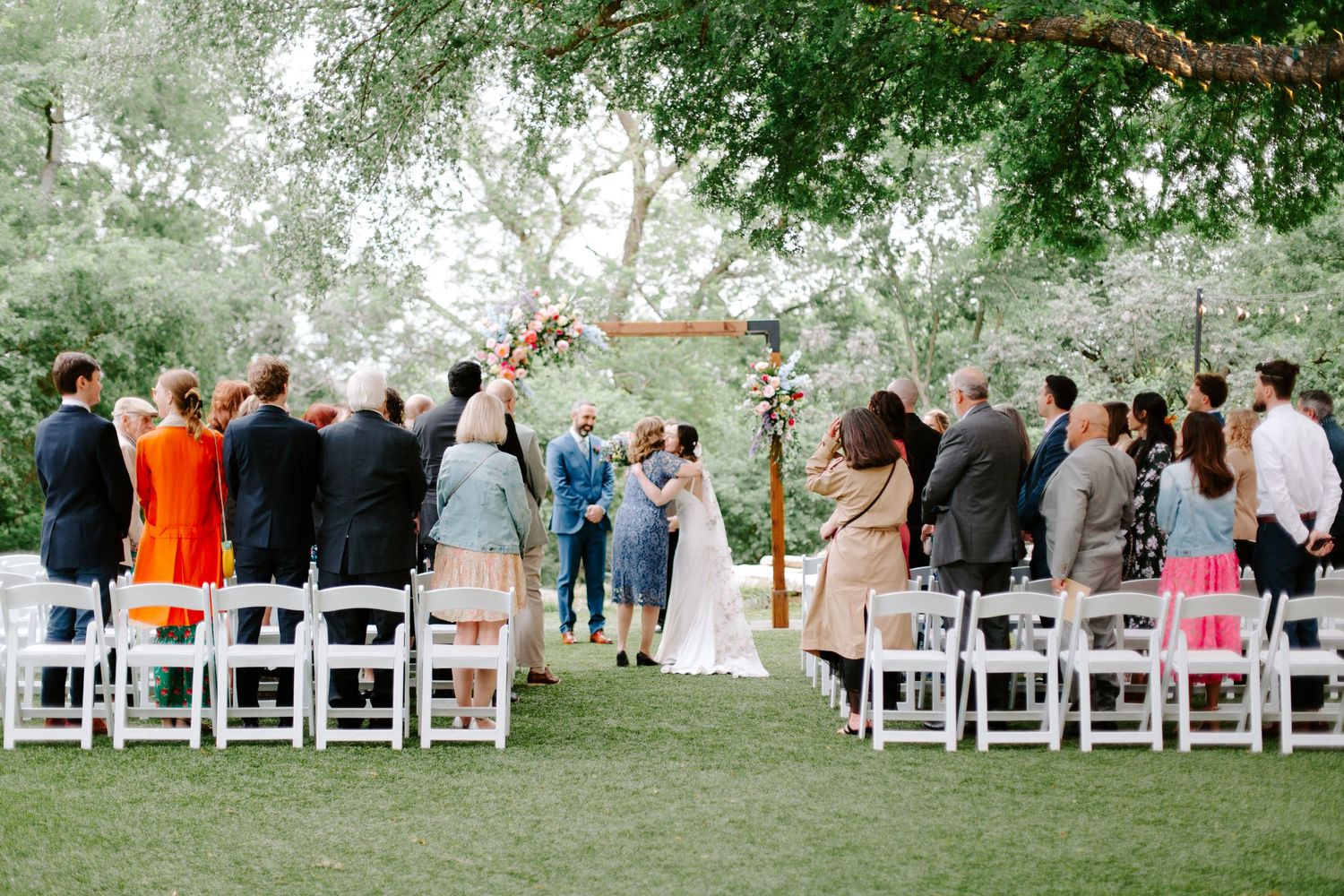 Wedding ceremony taking place outdoors with white chairs set up on grass under tree canopy during daytime event.