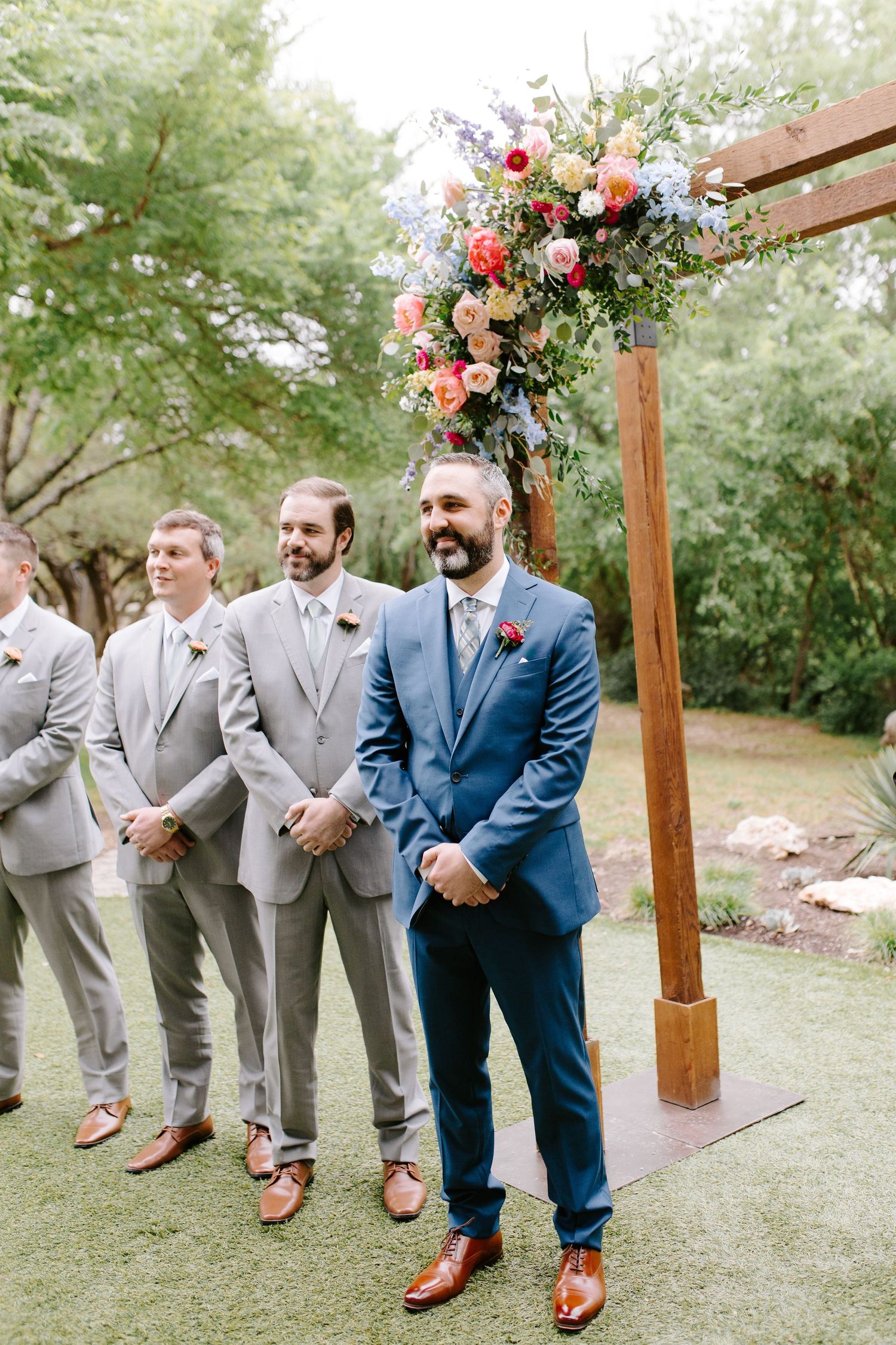 Groomsmen showing off their stylish brown leather shoes while standing on grass under a wooden arch with floral decor.