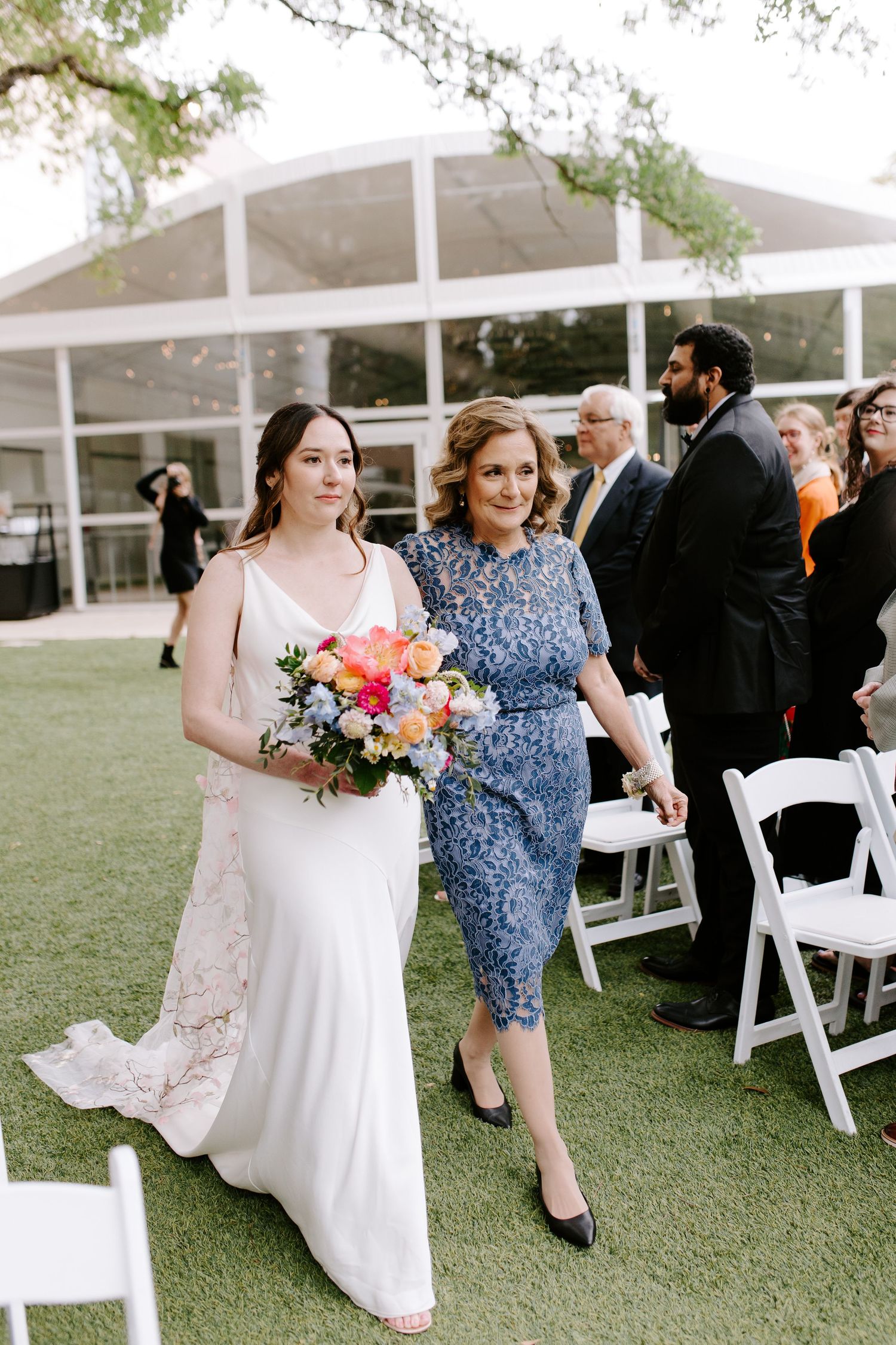 Two women walk together at an outdoor wedding ceremony, one in a white bridal gown holding flowers and another in blue dress.