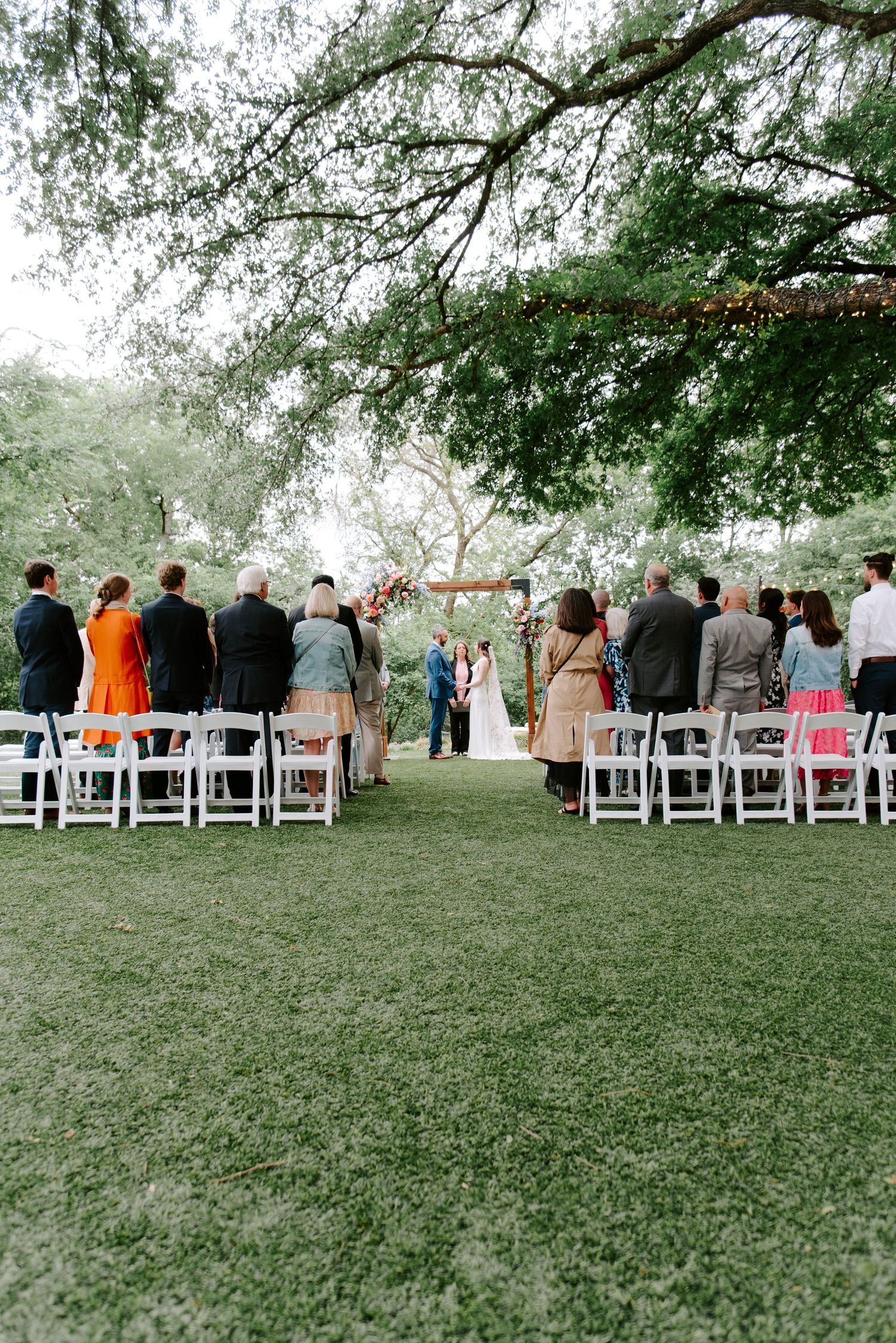 An outdoor wedding ceremony setup with white chairs under sprawling tree branches and string lights.