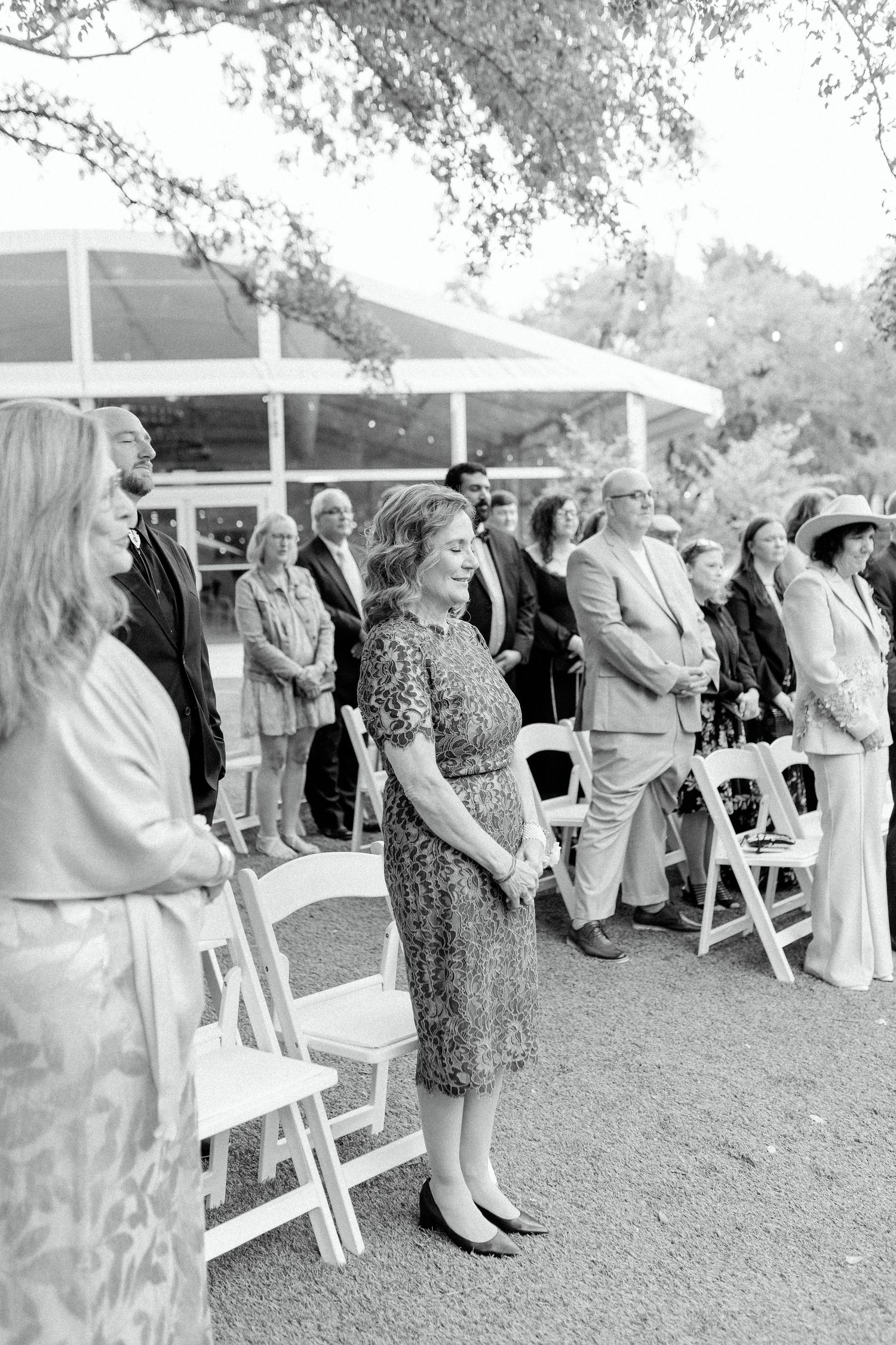 Black and white photo showing guests in formal attire standing and sitting at an outdoor event with white folding chairs.
