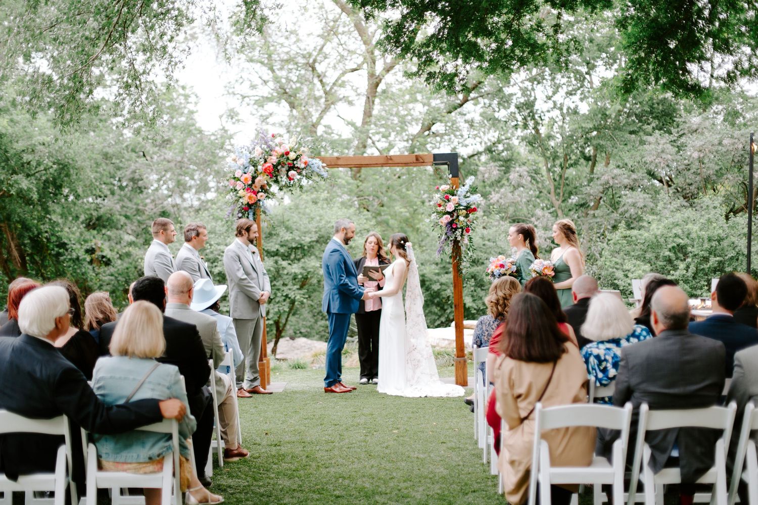 Wedding ceremony under a floral arch with guests seated on white chairs in a garden setting.