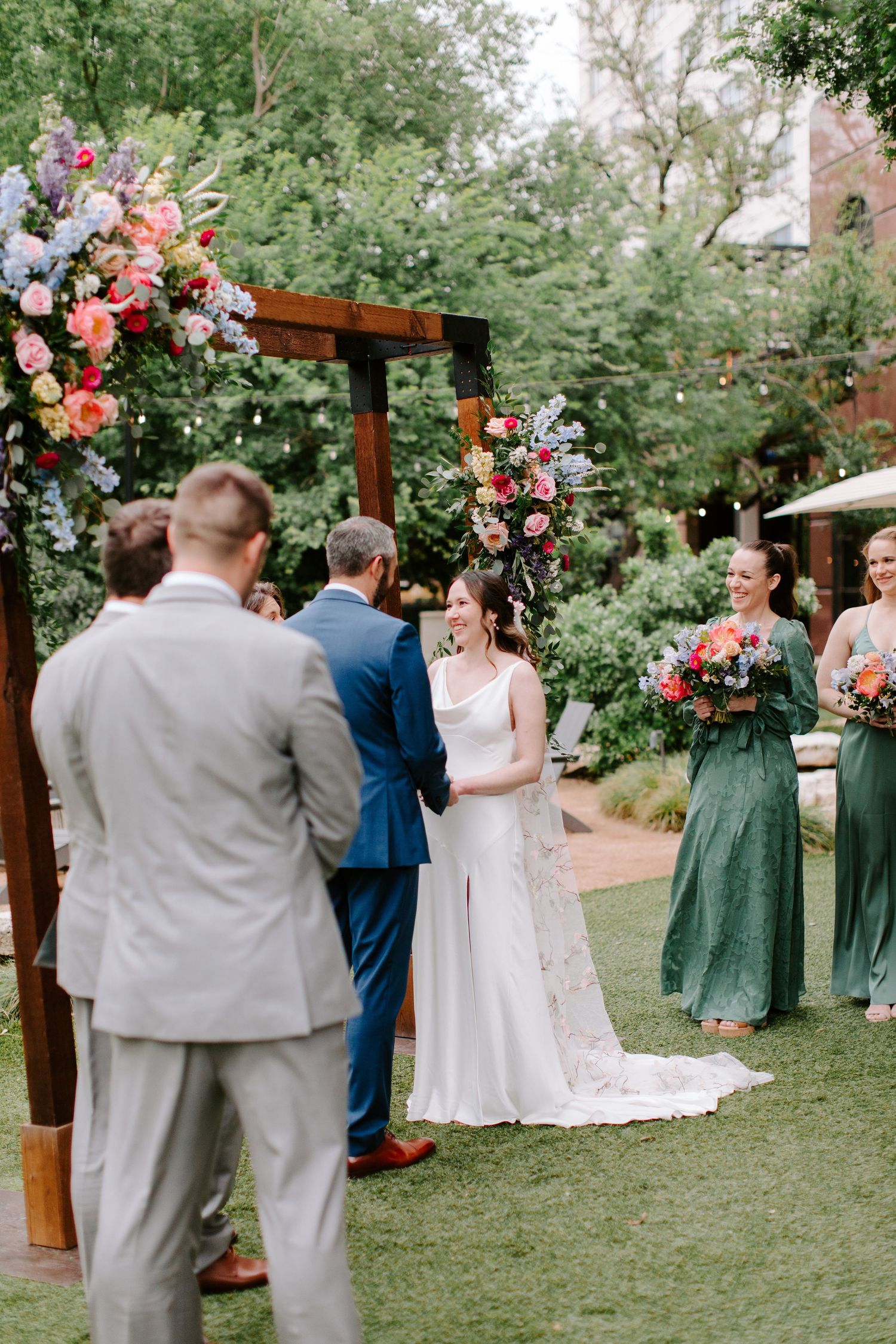 A wedding ceremony takes place under a wooden arch adorned with pink and purple flowers in a lush garden setting.