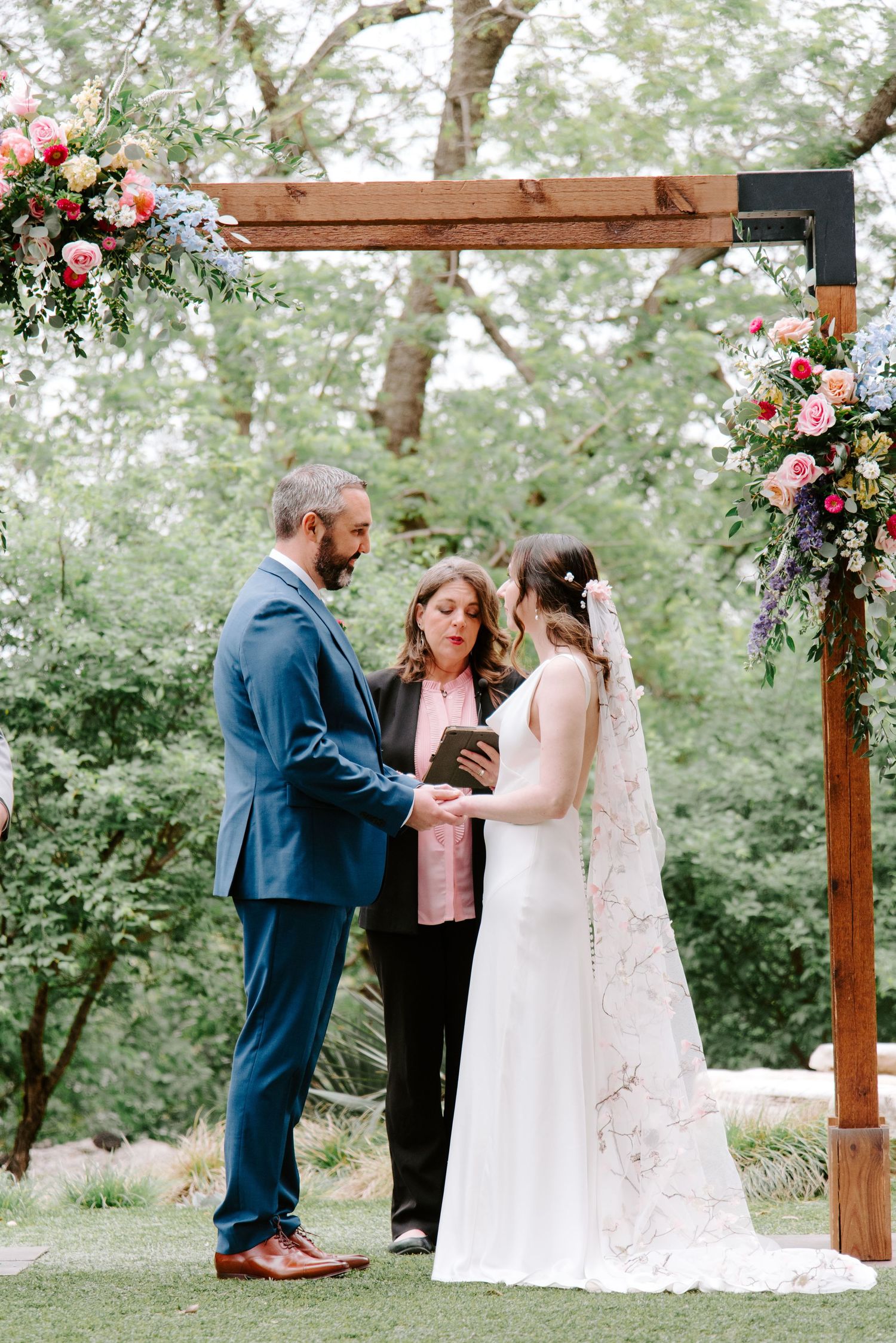 Outdoor wedding ceremony under a wooden arch decorated with pink and white floral arrangements in a garden setting.
