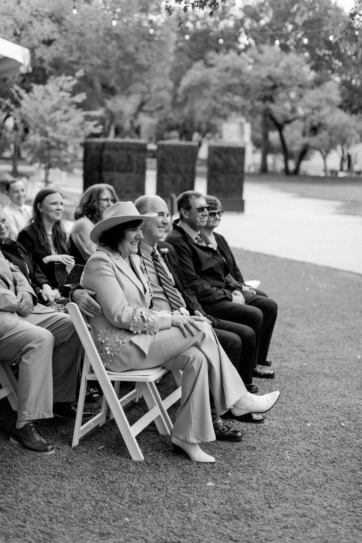 A group of guests sitting on white folding chairs outdoors in a garden or park setting, captured in black and white.