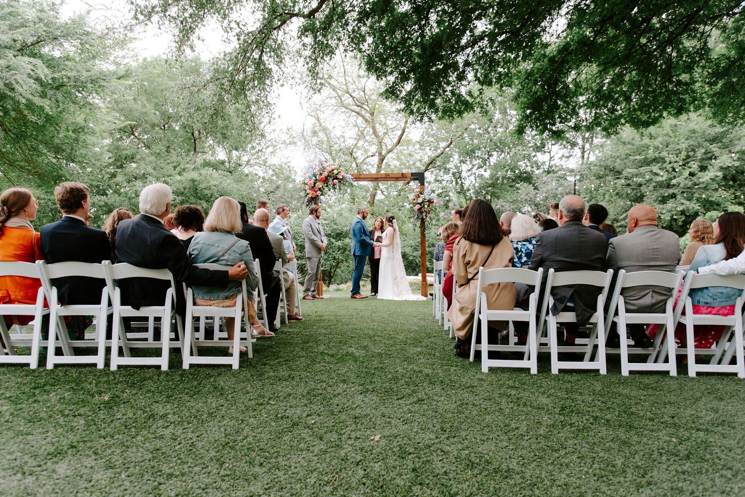 Outdoor wedding ceremony with guests seated in white chairs along a grass aisle leading to a wooden arch.