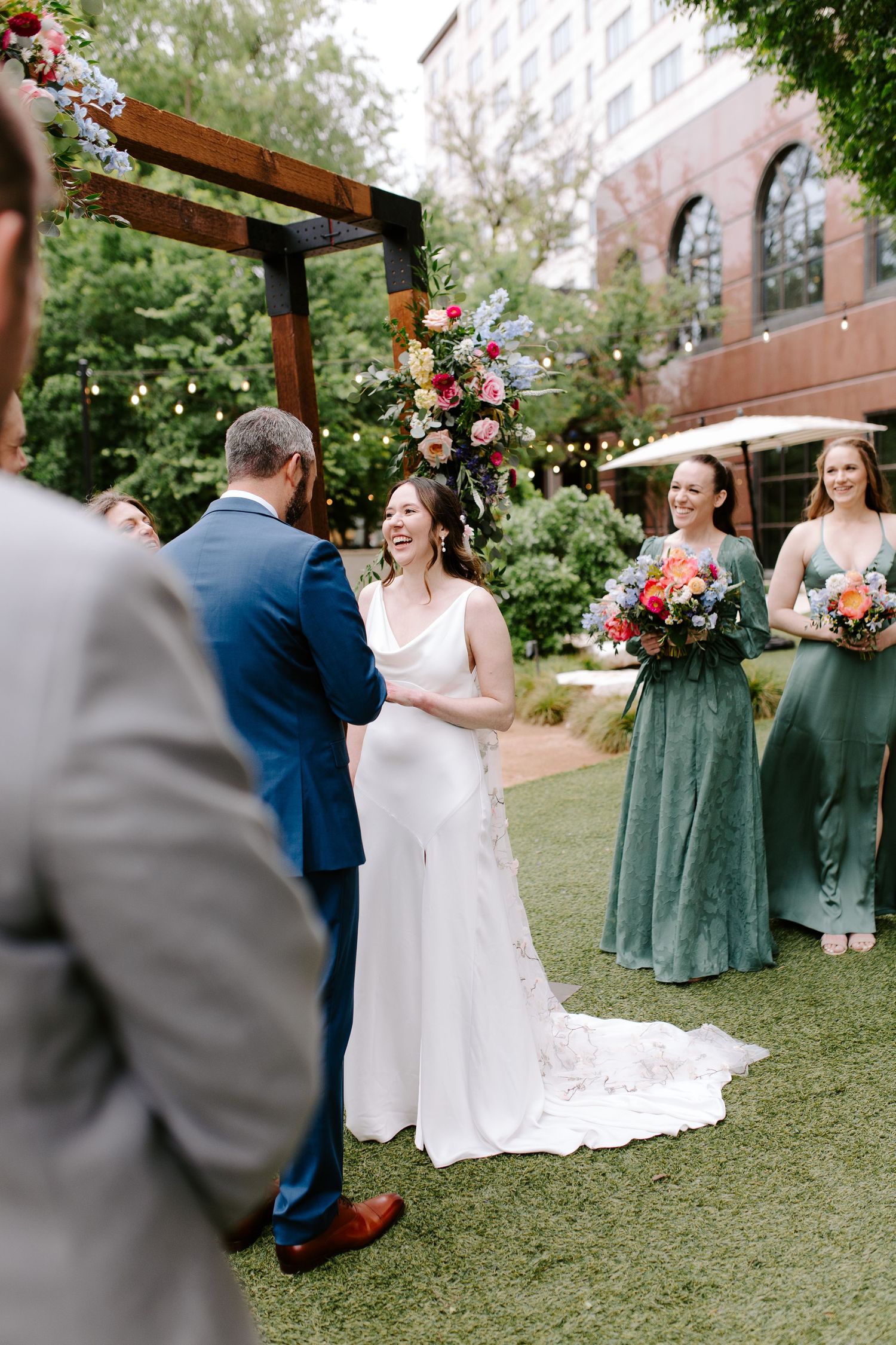 Outdoor wedding ceremony with bridesmaids in sage green dresses standing beside a floral-decorated wooden arch.
