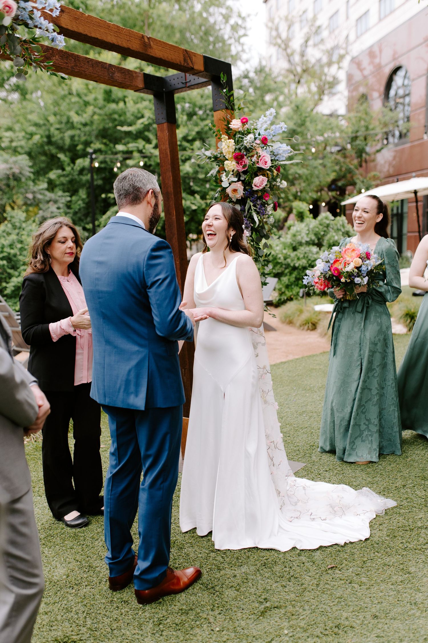 Wedding ceremony under a wooden arbor decorated with pink and white floral arrangements in an outdoor garden setting.