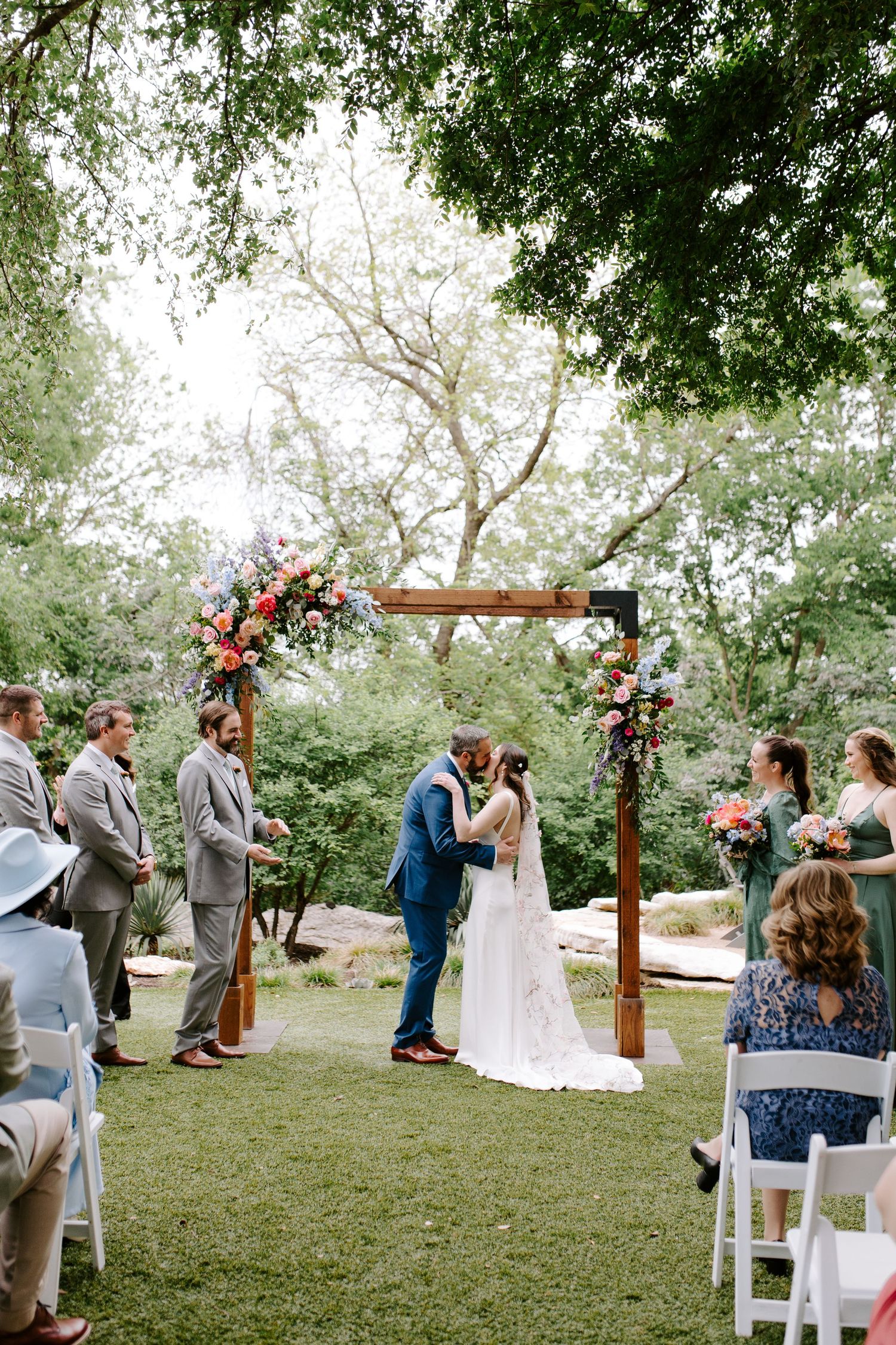 Outdoor wedding ceremony under wooden arch decorated with pink and red flowers surrounded by lush greenery.