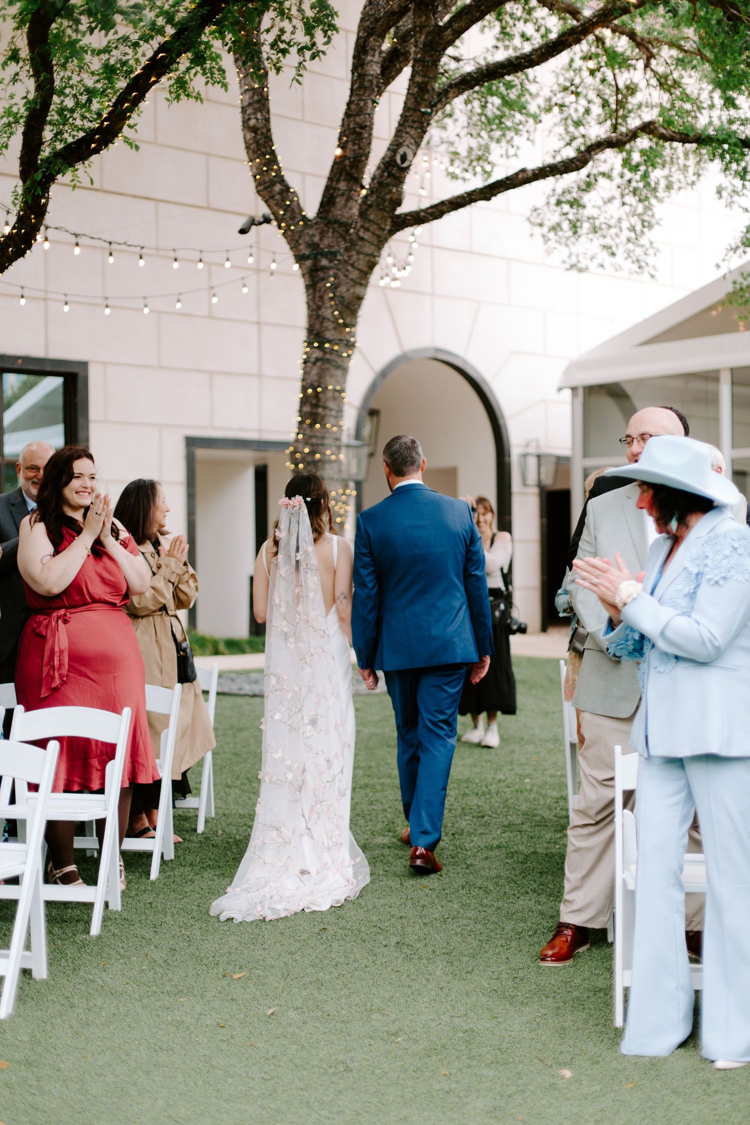 Wedding guests watch as a couple walks down the aisle decorated with string lights and trees.