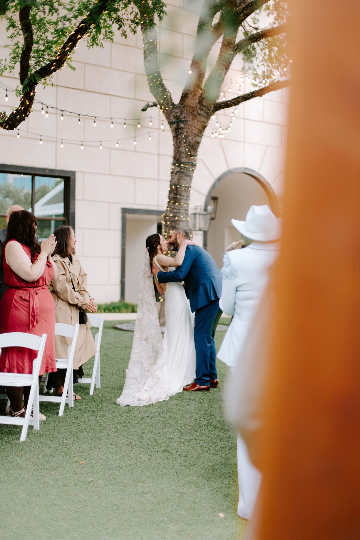 Outdoor wedding ceremony under a large tree with string lights and white chairs on green turf.