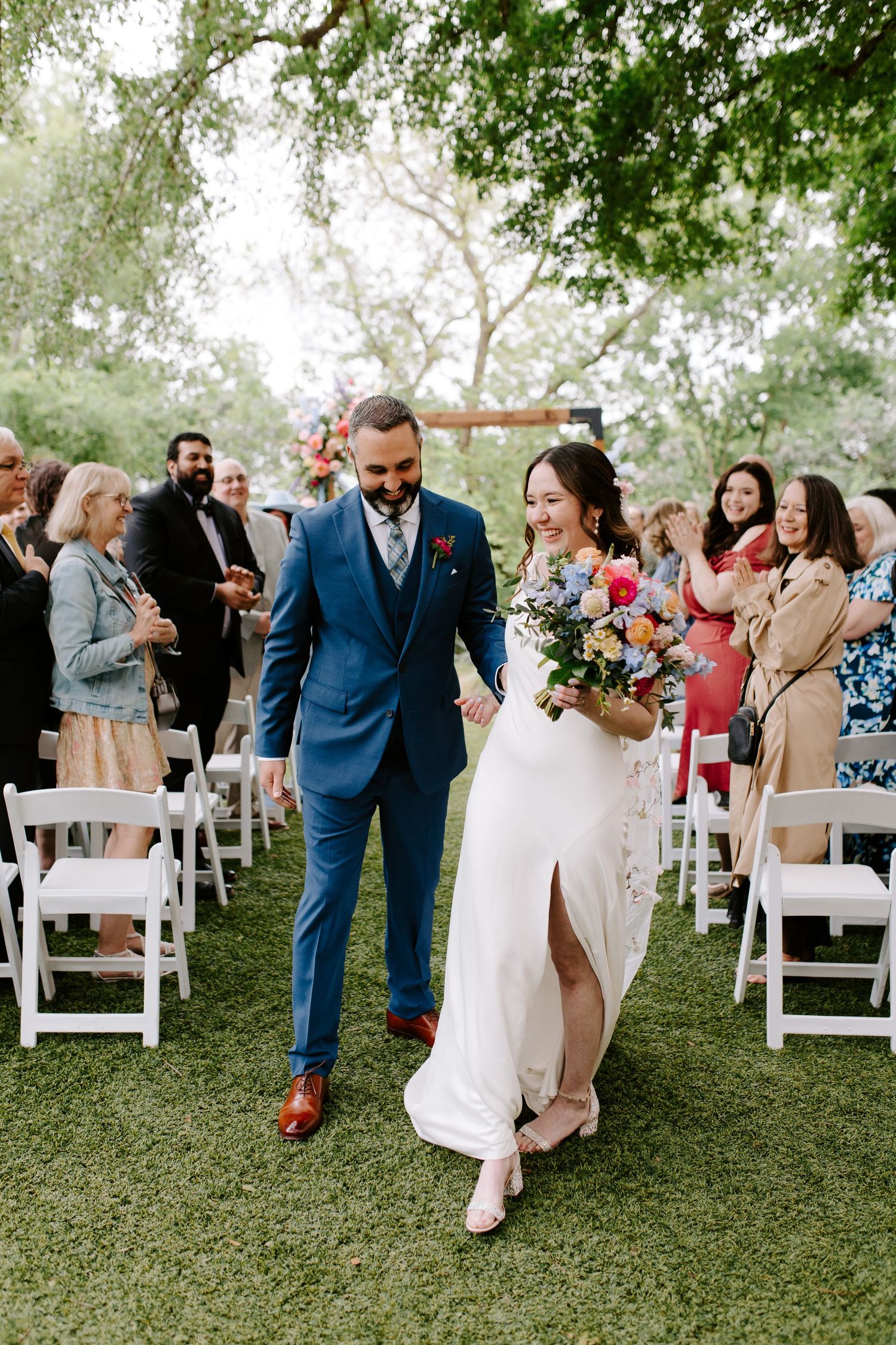 Newlyweds walk down the aisle while wedding guests celebrate on white chairs under trees.